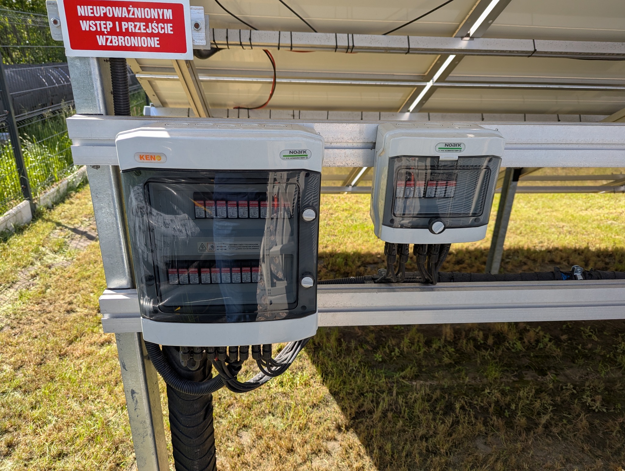 Dwa białe rozdzielacze elektryczne KENO i NOARK zamontowane pod panelami słonecznymi, z widocznymi kablami i tabliczką ostrzegawczą.