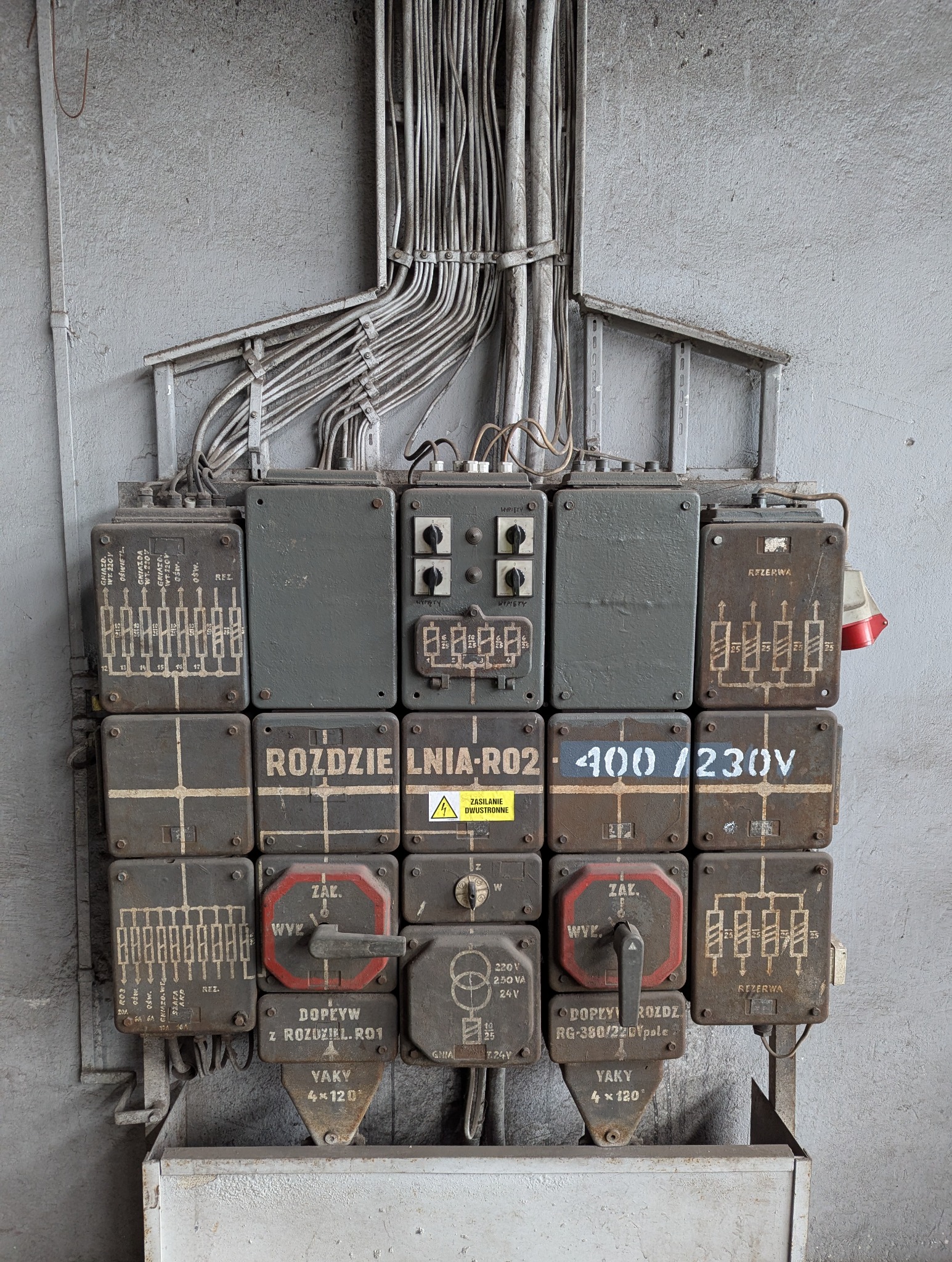 Stara, przemysłowa rozdzielnia elektryczna z przewodami na szarej, betonowej ścianie. Widoczne oznaczenia, schematy i przełączniki. Charakterystyczny, techniczny detal.