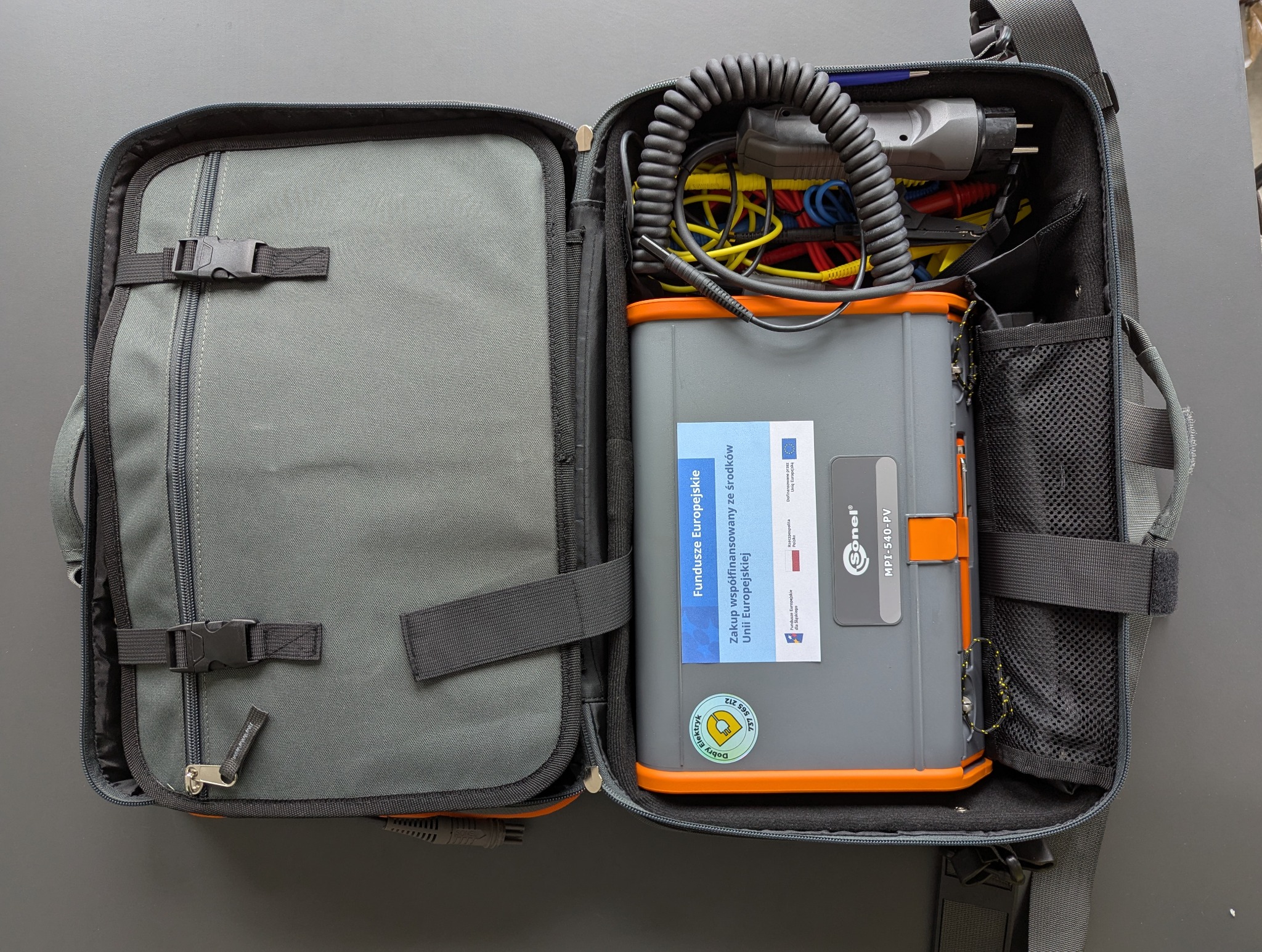 Otwarta torba instalatora z miernikiem Sonel MPI-540-PV, przewodami i naklejką 'Dobry Elektryk'. Widoczna etykieta 'Fundusze Europejskie'.