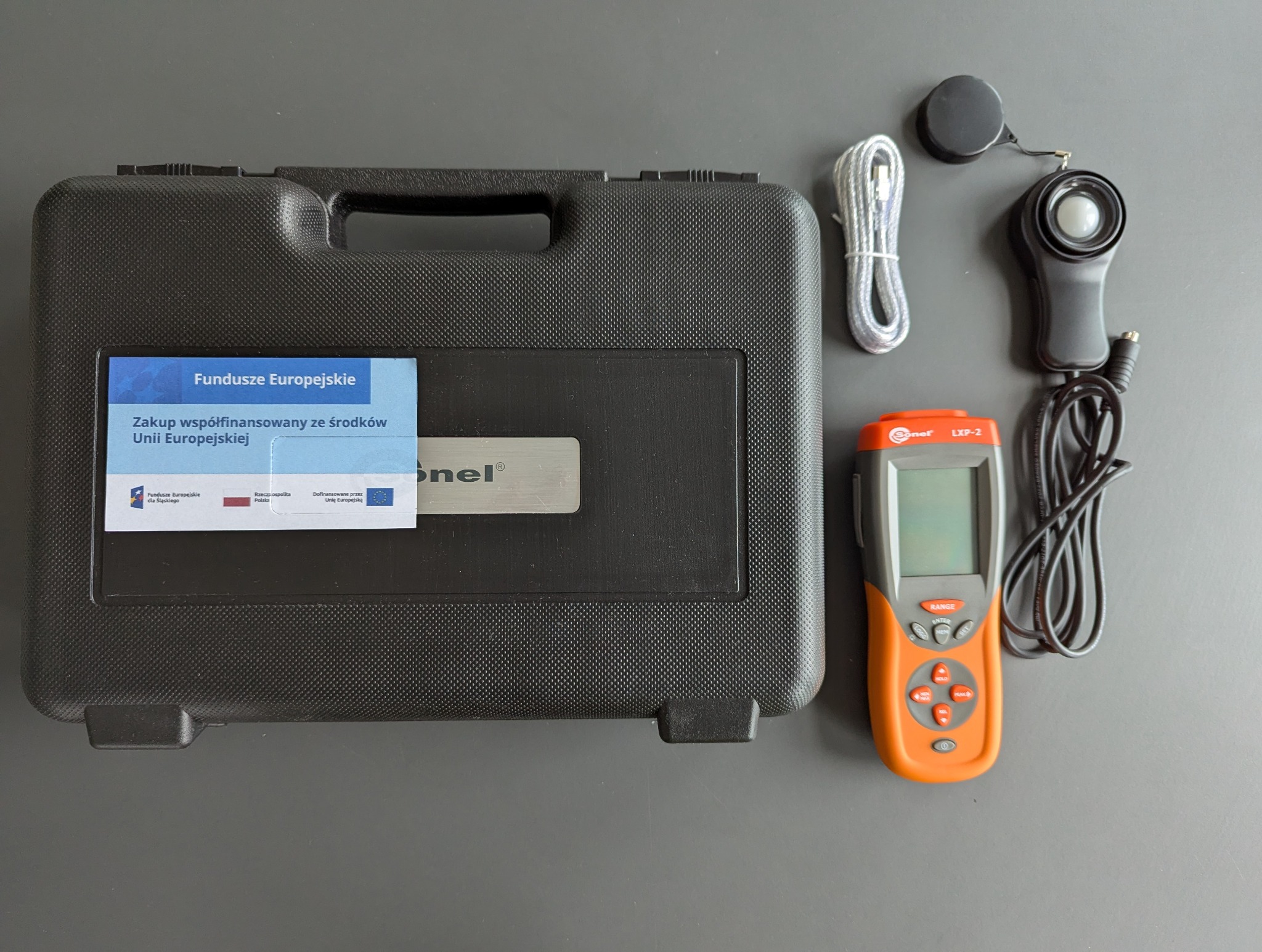 Luksometr Sonel LXP-2 w pomarańczowej obudowie, kabel, czujnik i czarna walizka z naklejką Funduszy Europejskich na szarym tle. Sprzęt do pomiarów elektrycznych.