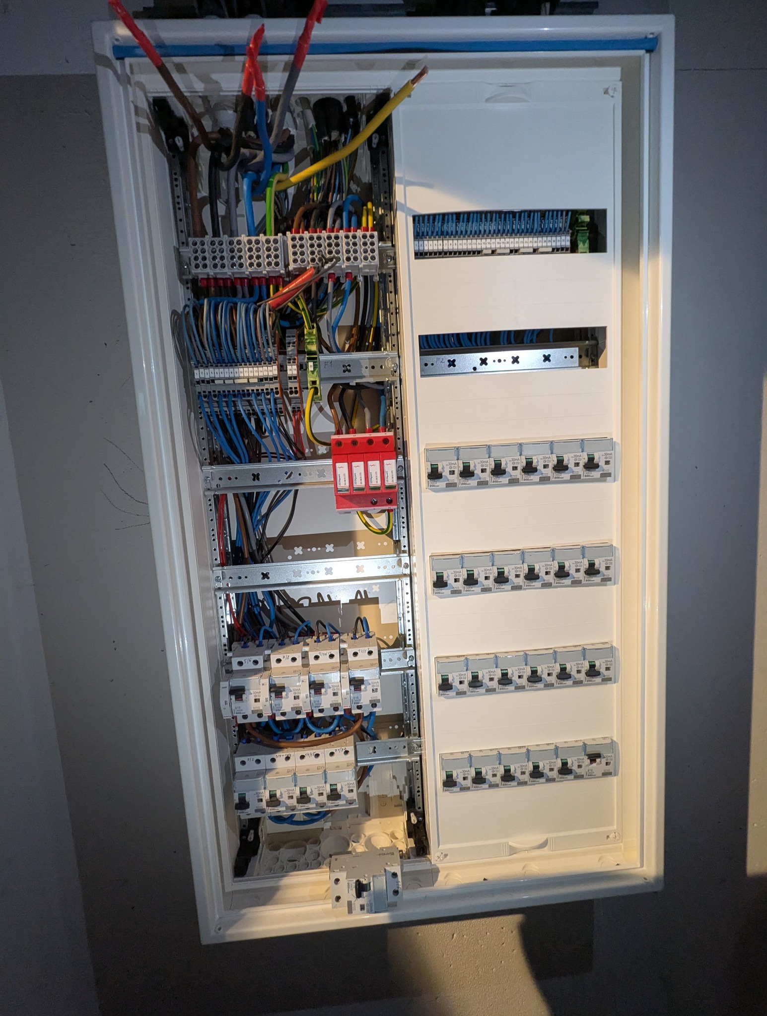 Rozdzielnica elektryczna z widocznymi przewodami, bezpiecznikami i zabezpieczeniami, zamontowana na szarej ścianie. Widoczne kable w różnych kolorach.