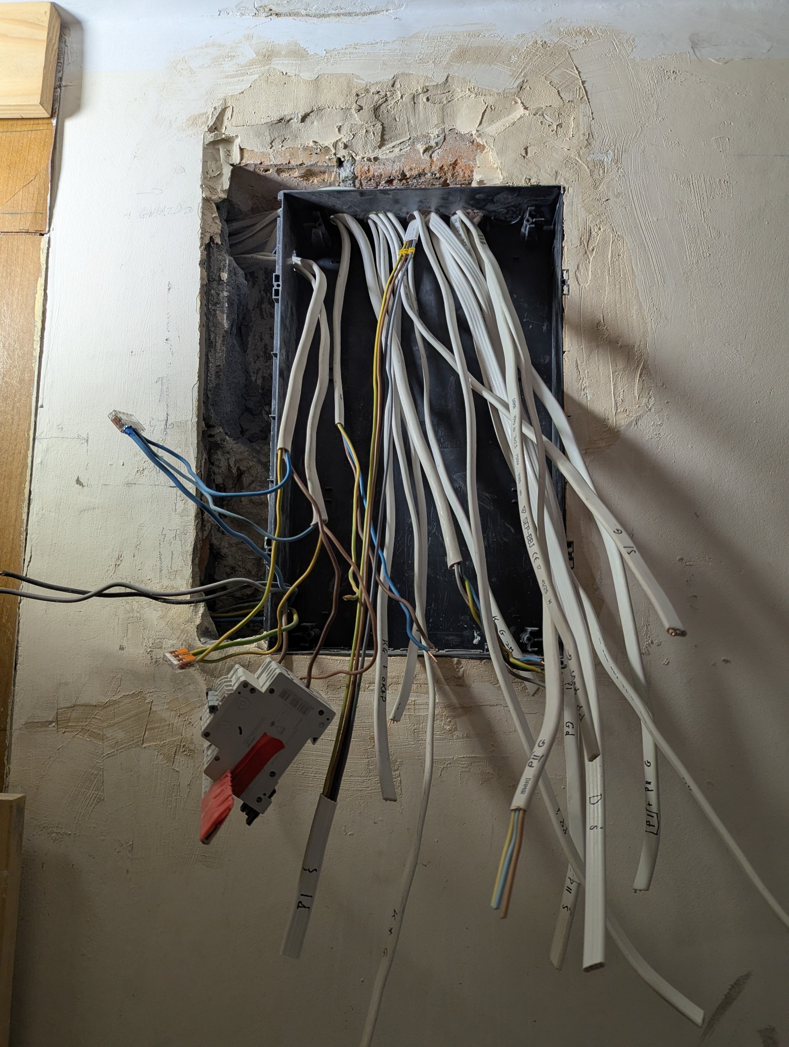 Otwarta puszka elektryczna w ścianie z wystającymi przewodami oznaczonymi napisami, widoczne elementy instalacji i surowy stan wykończenia ściany.