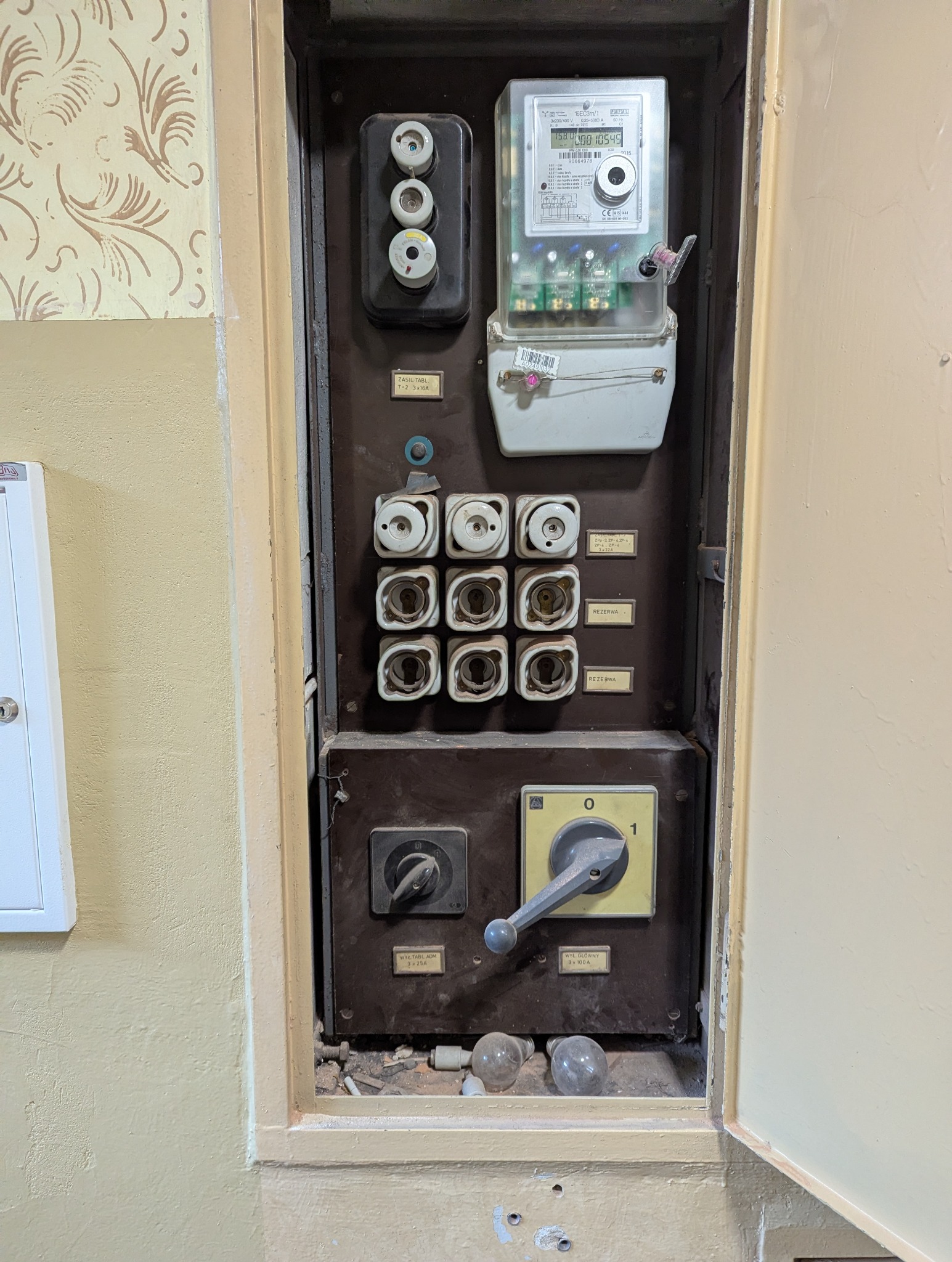 Otwarta, stara skrzynka elektryczna z bezpiecznikami, licznikiem i przełącznikami wewnątrz, umieszczona na ścianie z wzorzystą tapetą i pomalowaną farbą.