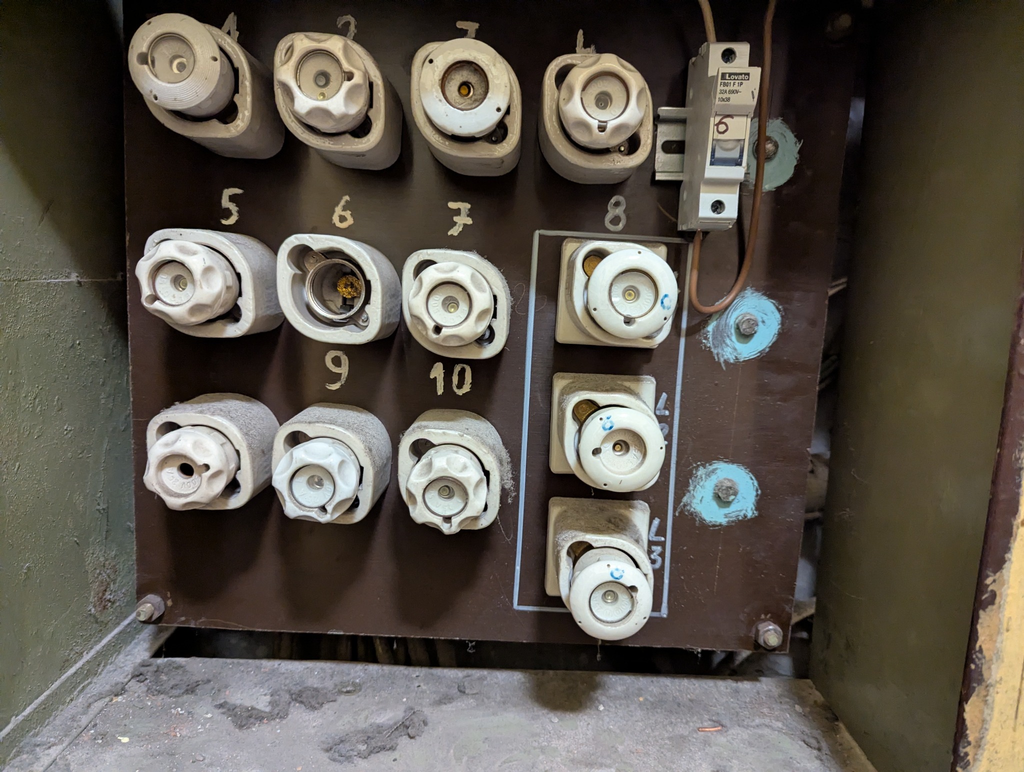 Tablica bezpiecznikowa z ceramicznymi bezpiecznikami topikowymi i wyłącznikiem nadprądowym, numery od 5 do 10, widoczne ślady użytkowania i zabrudzenia.