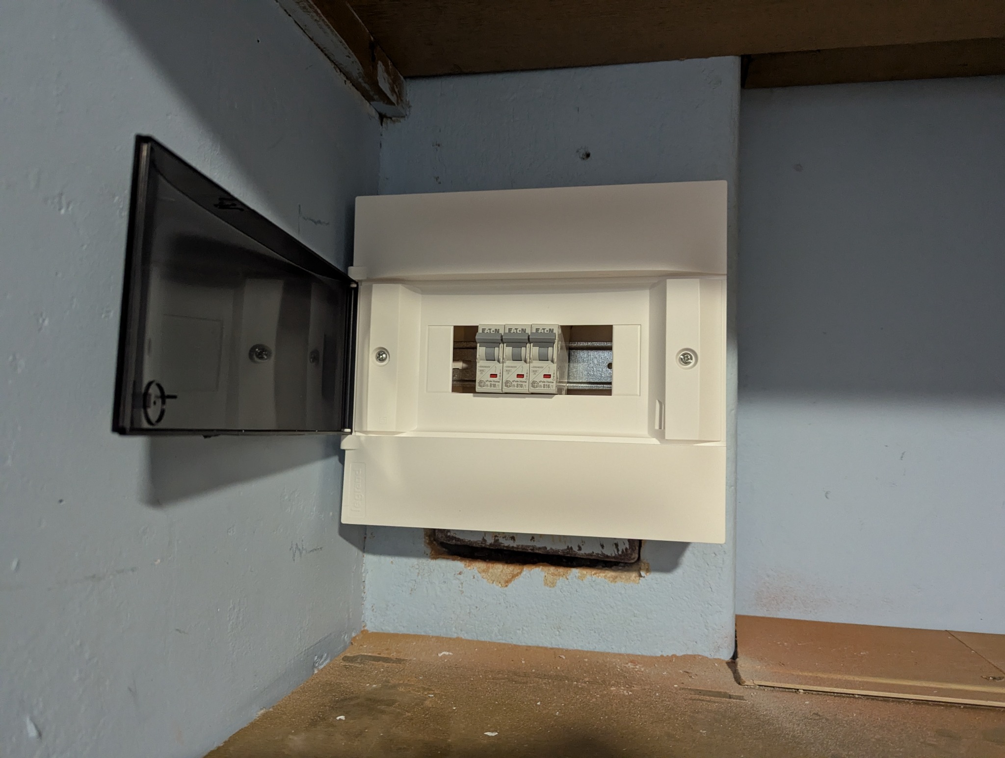 Biała skrzynka rozdzielcza z bezpiecznikami Eaton, zamontowana na niebieskiej ścianie. Drzwiczki skrzynki otwarte po lewej stronie, widoczne trzy bezpieczniki.