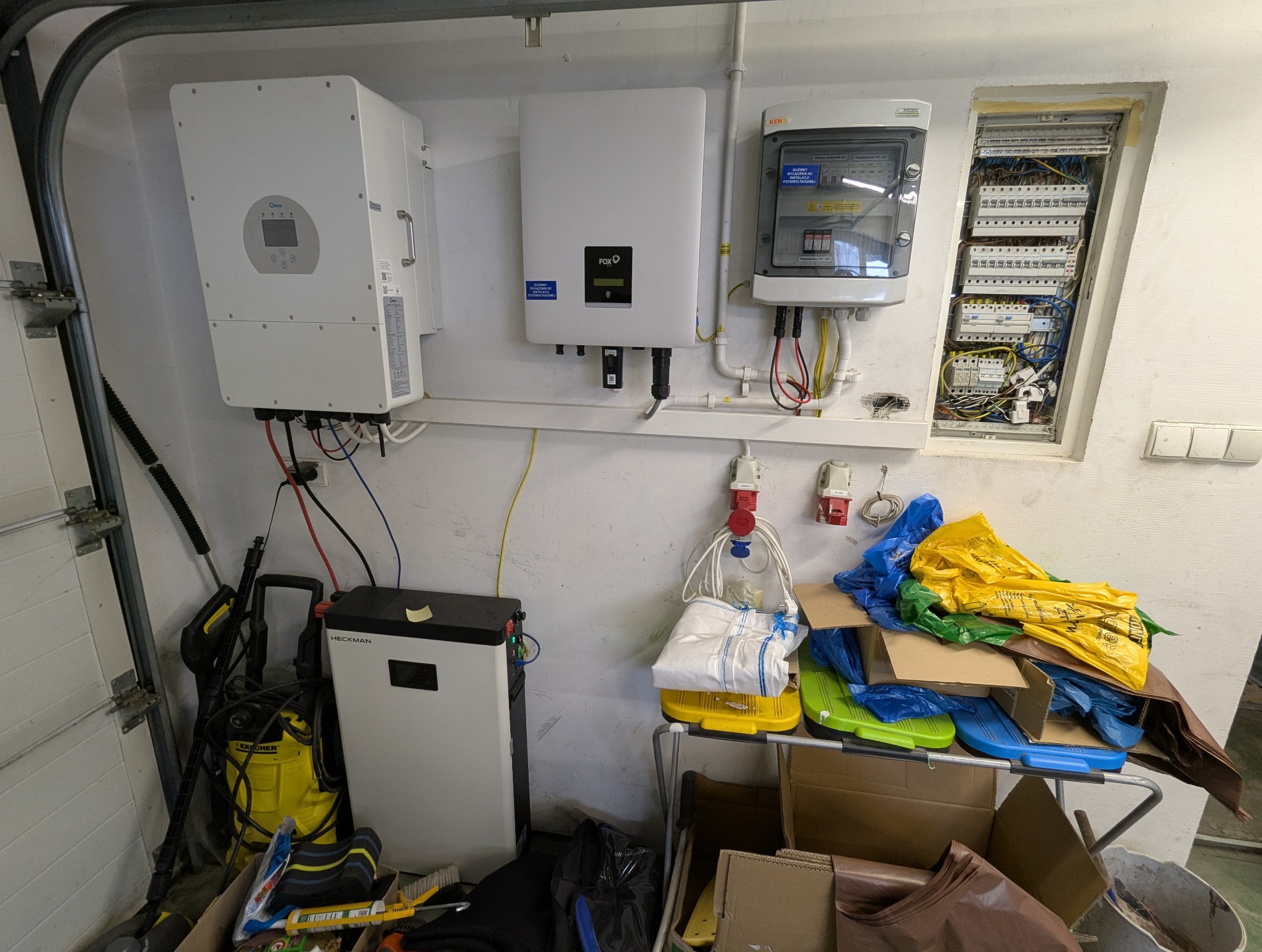 Instalacja fotowoltaiczna w garażu: inwertery Deye i Fox, akumulator Heckman, rozdzielnica elektryczna. Widoczne kable, przewody i narzędzia. Częściowy widok bramy garażowej.