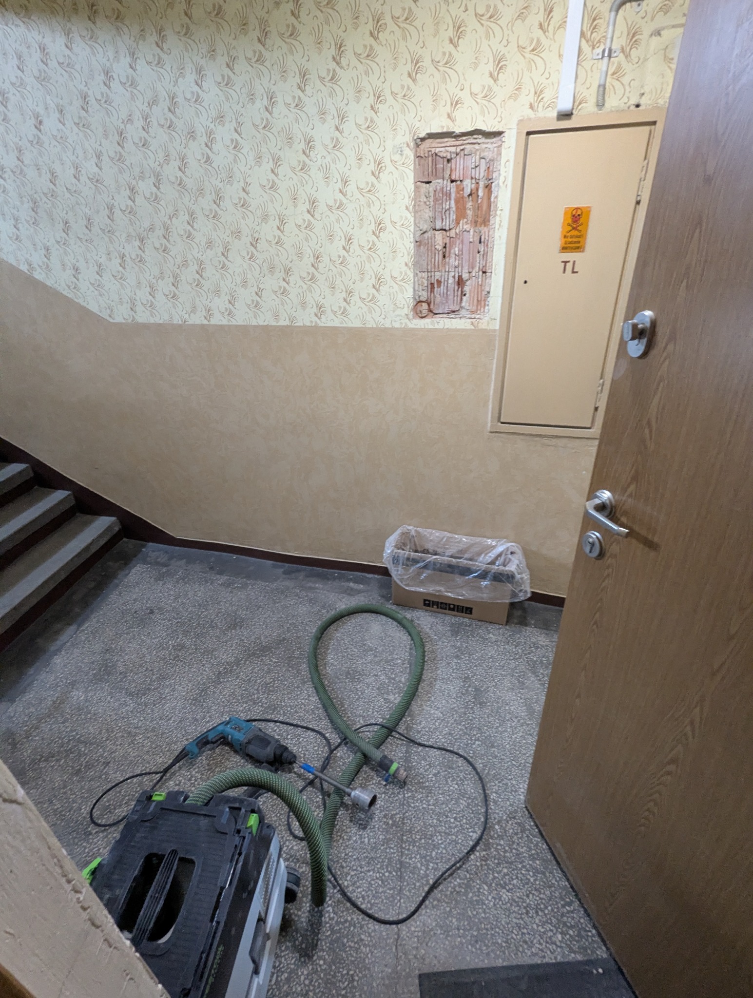 Otwór w ścianie z cegły na klatce schodowej, obok skrzynka elektryczna. W tle tapeta w kwiaty. Na podłodze wiertarka i odkurzacz przemysłowy.