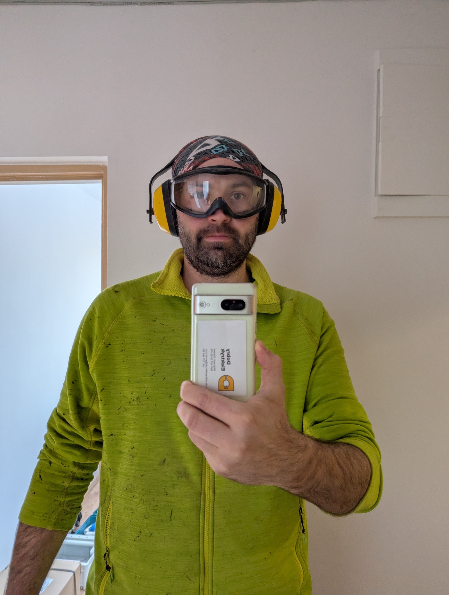 Elektryk w okularach ochronnych i słuchawkach, robiący selfie telefonem z logo firmy z Gliwic. Widoczne ślady pracy na ubraniu.