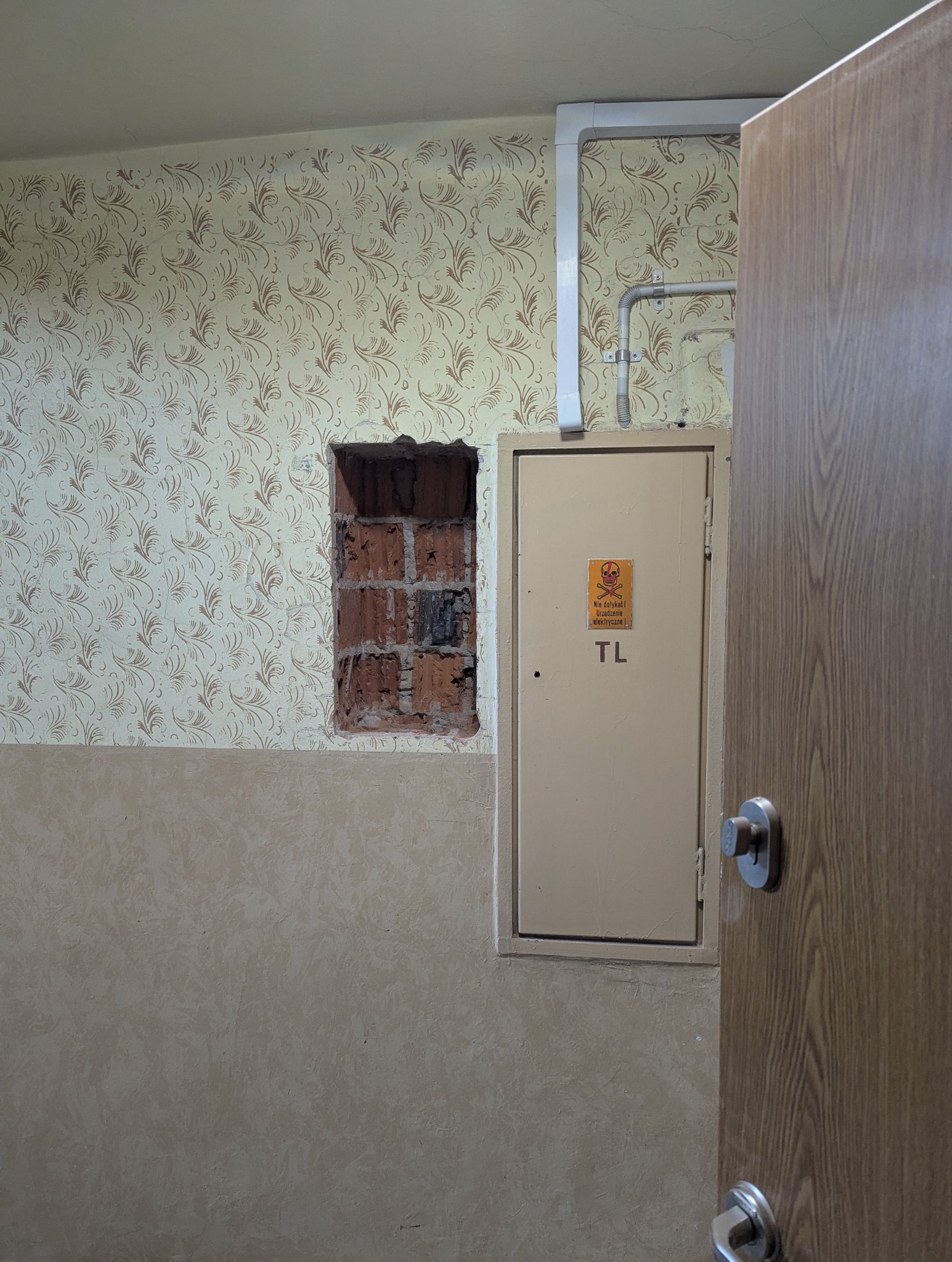 Otwarta szafka elektryczna z ostrzeżeniem na ścianie z wzorzystą tapetą i odsłoniętą cegłą, widoczne przewody w białej listwie.