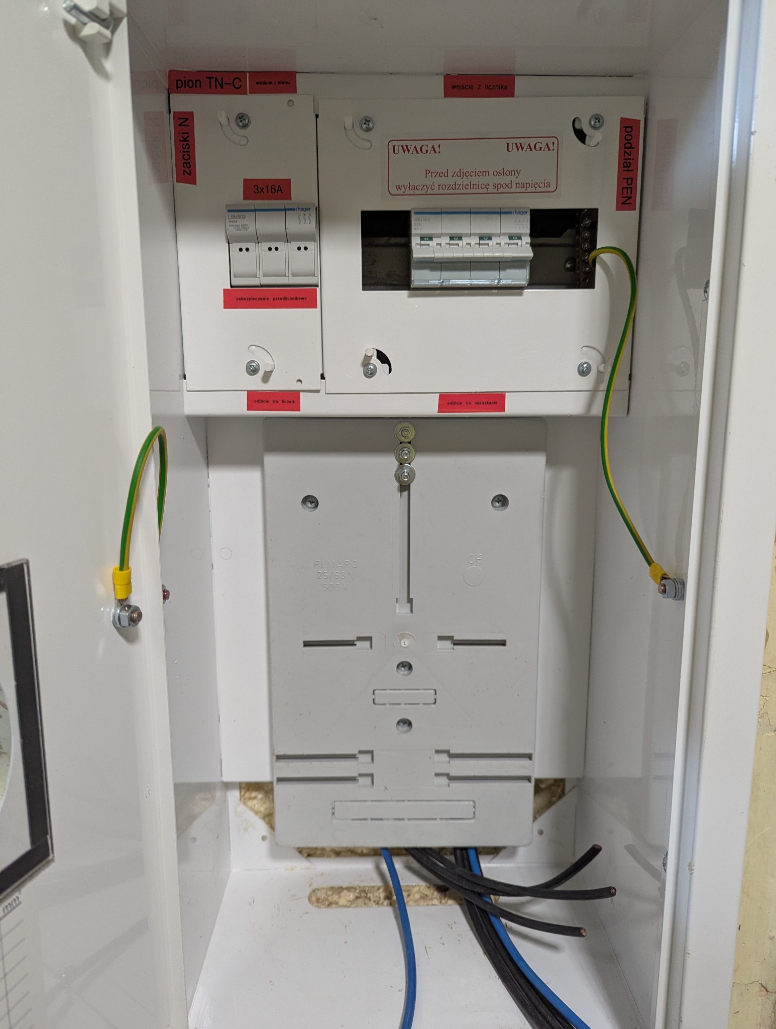 Otwarta skrzynka elektryczna z bezpiecznikami, przewodami i oznaczeniami. Widoczne zabezpieczenia przedlicznikowe, podział PEN i tabliczka ostrzegawcza.