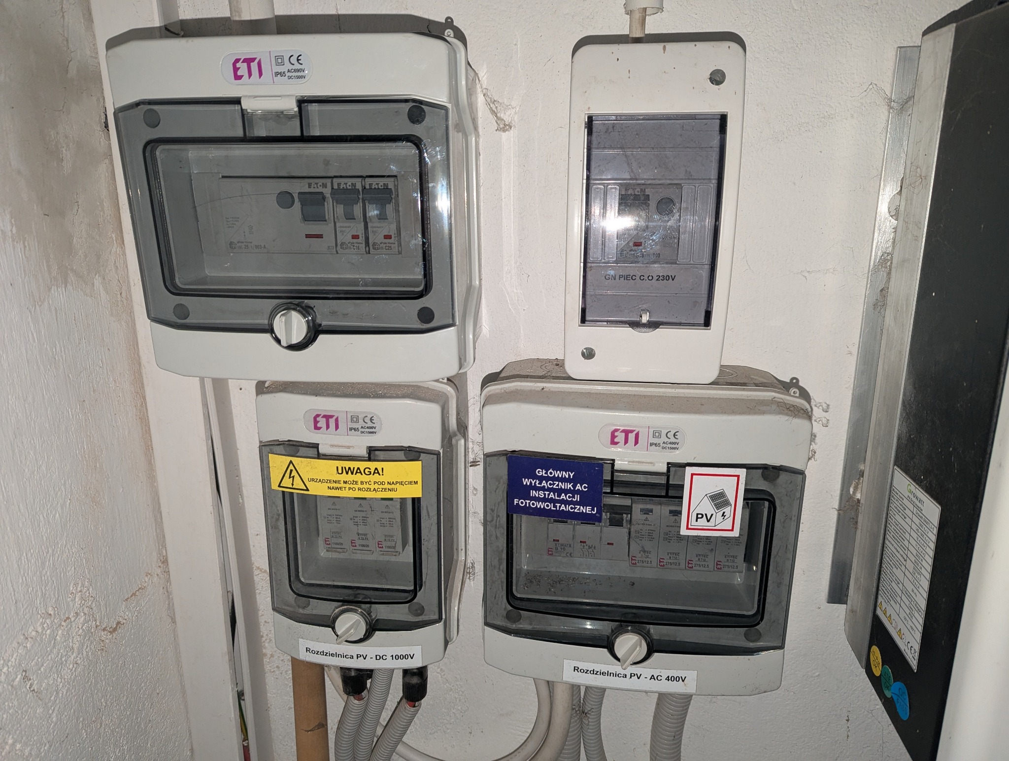 Rozdzielnice elektryczne ETI: DC 1000V, AC 400V, GN PIEC C.O 230V, z oznaczeniami ostrzegawczymi i informacyjnymi, zamontowane na ścianie. Widoczne kable.