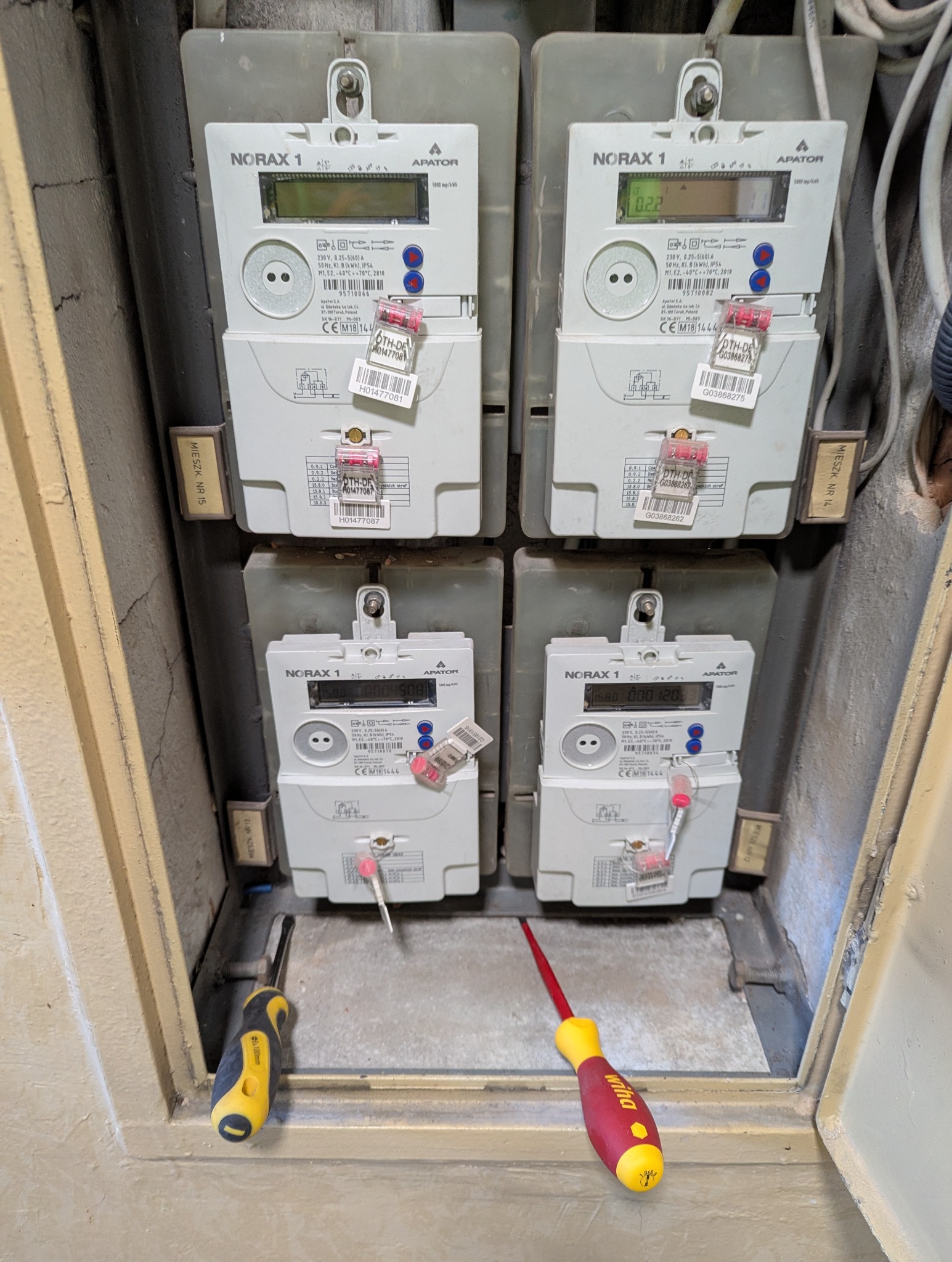 Cztery liczniki elektryczne Norax 1 w szarej skrzynce, widoczne narzędzia: śrubokręty z żółto-czarną i czerwono-żółtą rękojeścią. Pomiar energii elektrycznej w mieszkaniu.