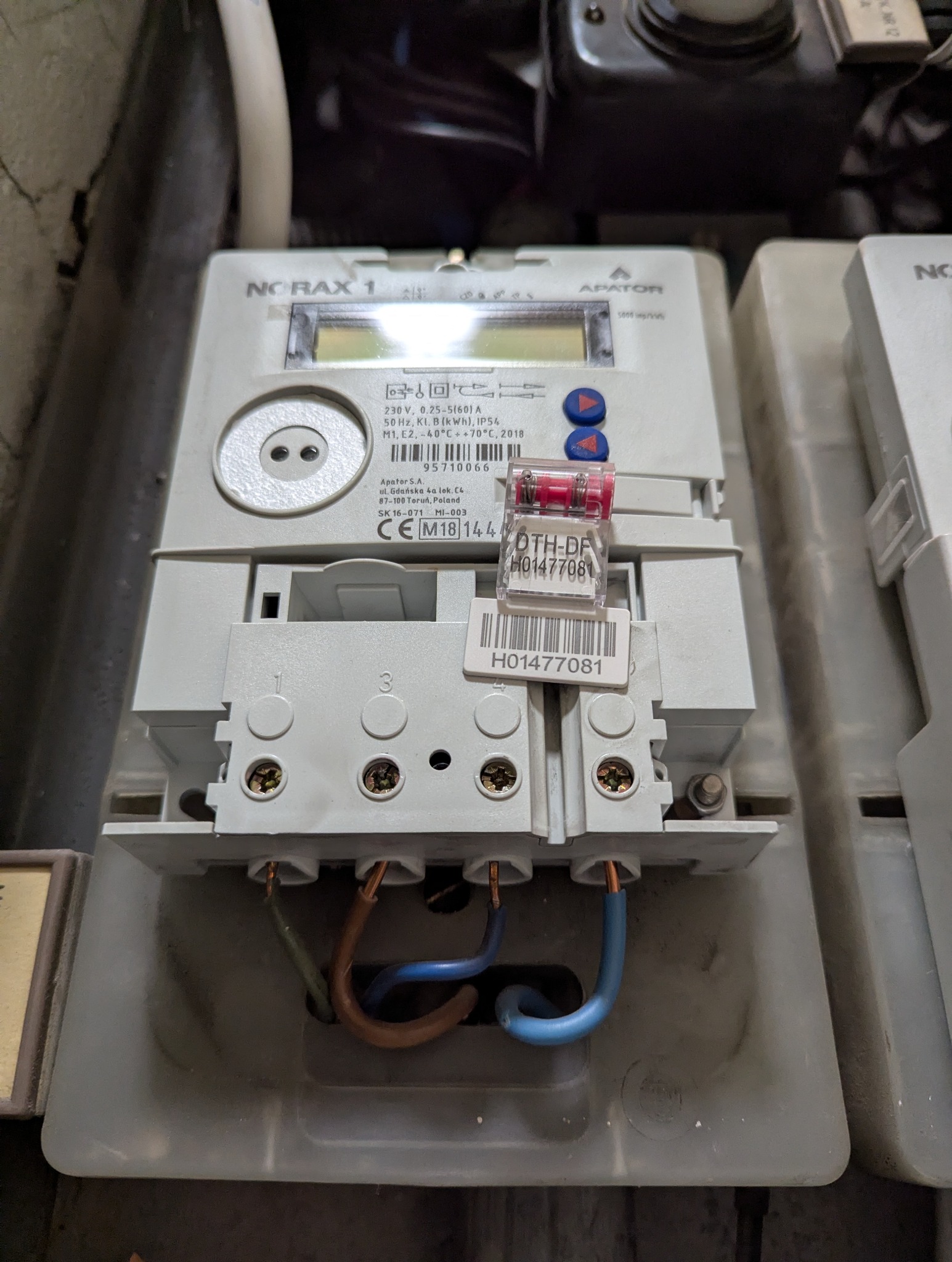 Szczegółowe ujęcie podłączonego licznika energii elektrycznej Norax 1 z widocznymi przewodami i plombą, zamontowanego w szarej obudowie.