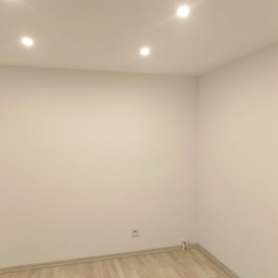 Paweł - Pomalowane na biało wnętrze pokoju z trzema punktowymi lampami sufitowymi i jasną podłogą. Widoczny fragment framugi drzwi po prawej stronie kadru.