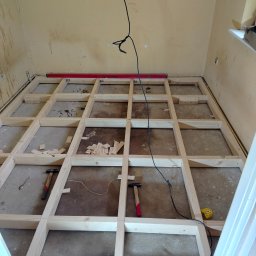 Paweł - Drewniana konstrukcja pod podłogę w trakcie montażu. Widoczne poziomica, młotki i miarka. Surowe ściany w tle. Etap przygotowania podłoża pod nową podłogę.
