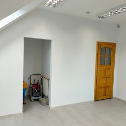 Paweł - Białe, świeżo pomalowane wnętrze biura z drewnianymi drzwiami i wnęką, w której stoi wózek z narzędziami malarskimi. Widoczne oświetlenie sufitowe.