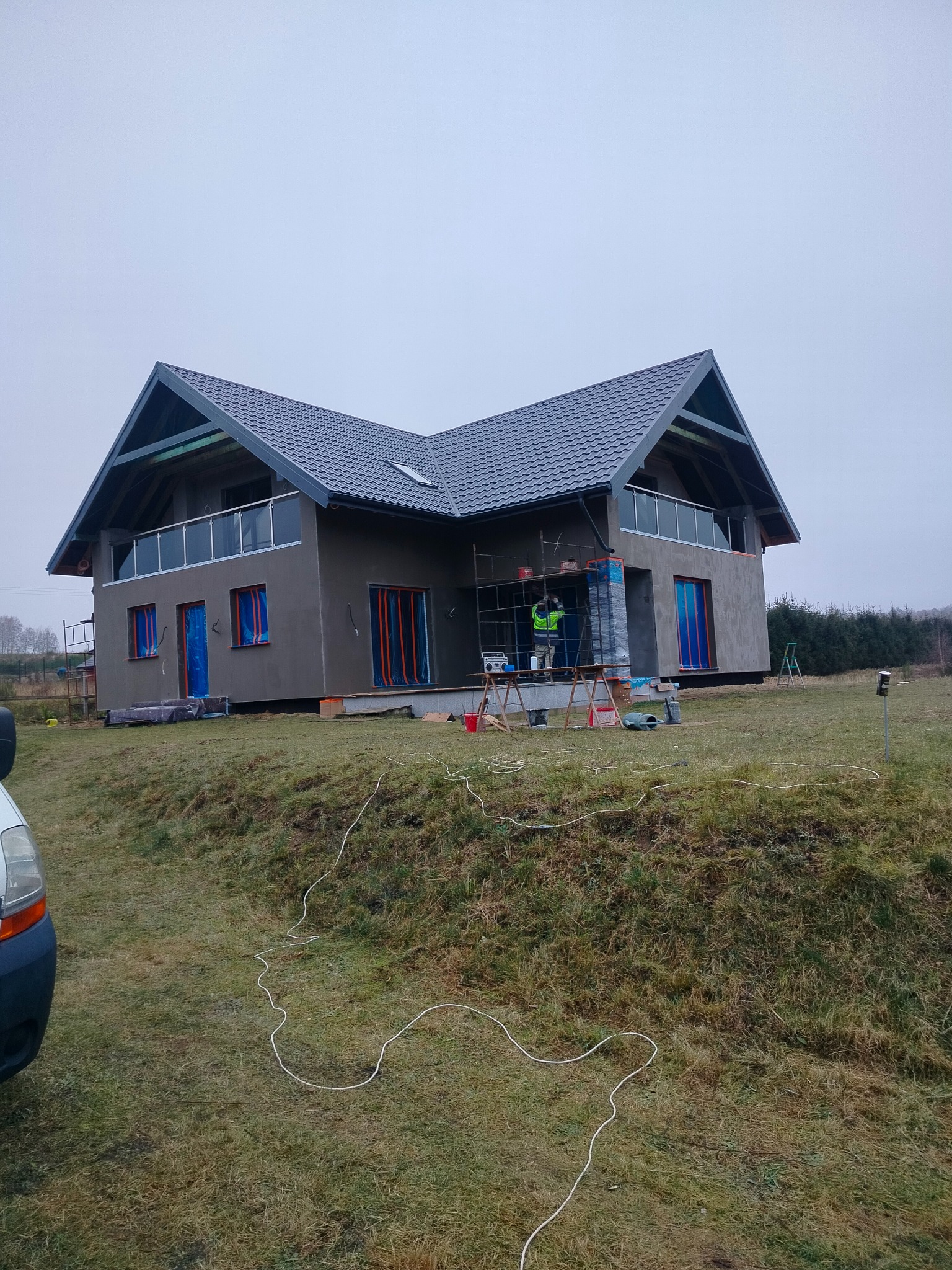 Dom jednorodzinny w trakcie prac wykończeniowych elewacji. Widoczne rusztowania, zabezpieczone okna i drzwi. Szara elewacja i dach z ciemnej dachówki.