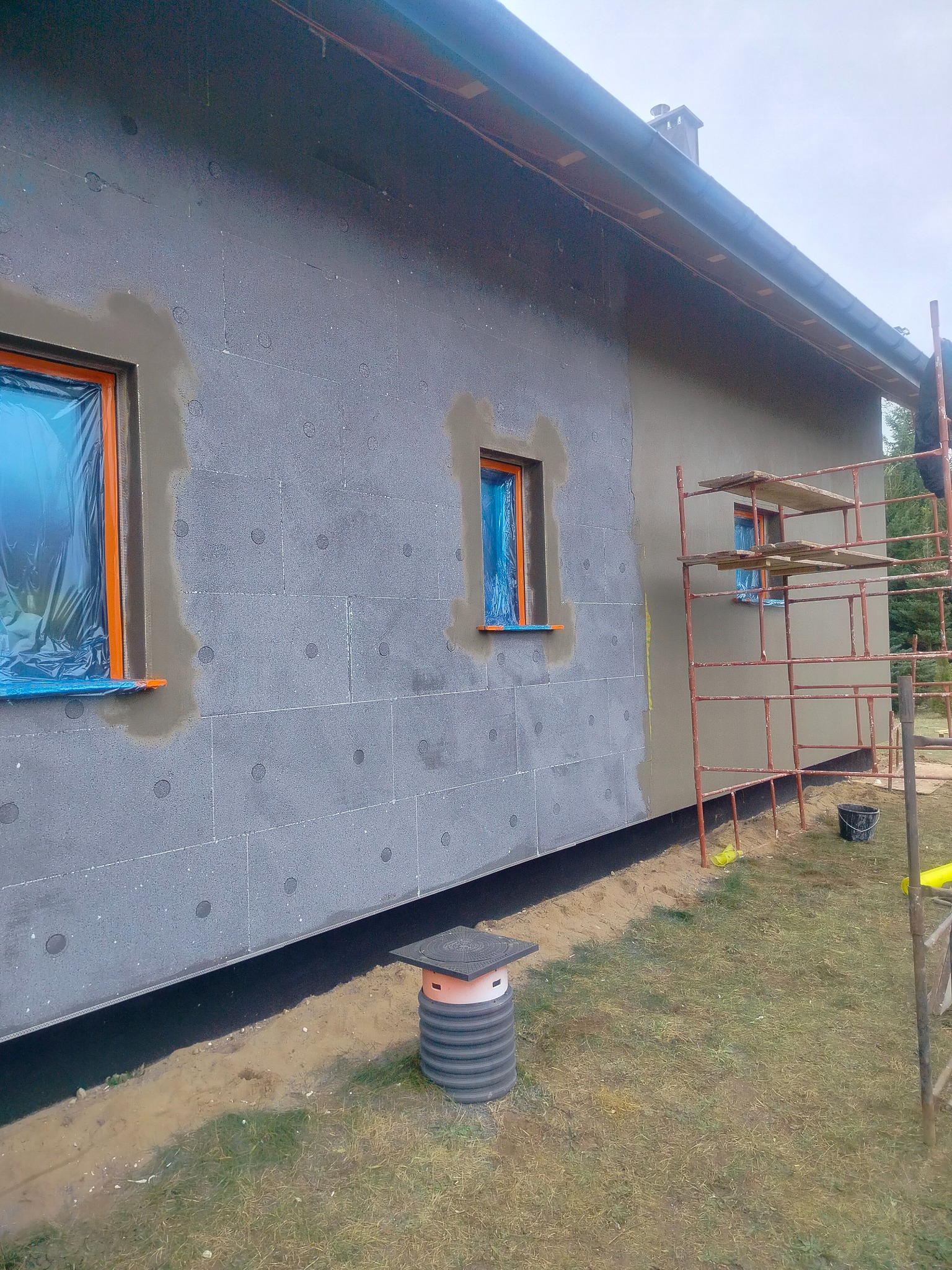 Ocieplanie elewacji domu: fragment ściany z szarym styropianem, okna zabezpieczone folią, część ściany otynkowana, rusztowanie. Widoczny etap prac.