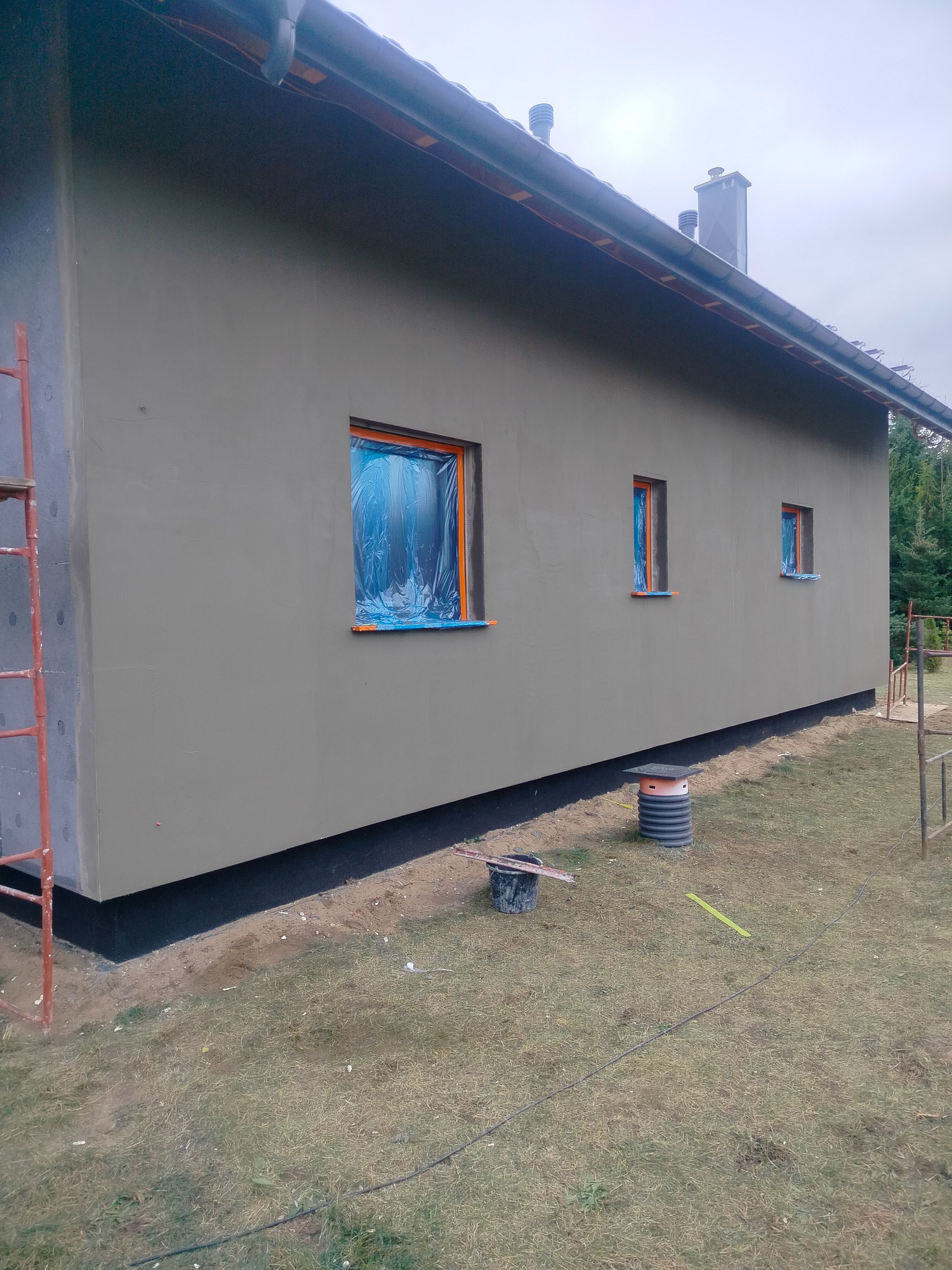 Elewacja domu po szpachlowaniu, okna zabezpieczone folią, widoczne narzędzia i rusztowanie. Świeżo nałożona szpachla w kolorze szarym.