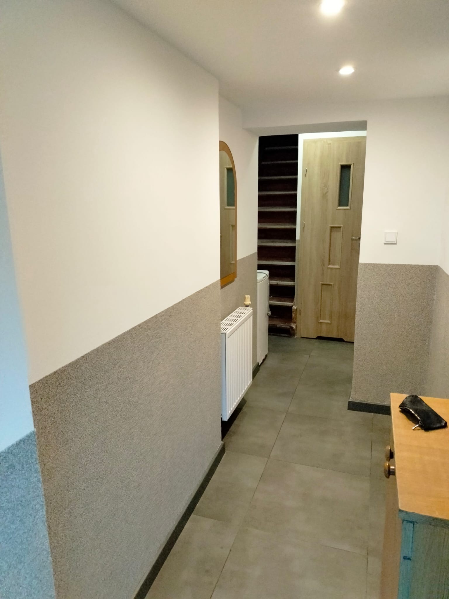 Wykończony korytarz z dwukolorowymi ścianami (biała góra, szary dół), szarą podłogą i drewnianymi schodami w tle. Widoczny grzejnik i fragment szafki.