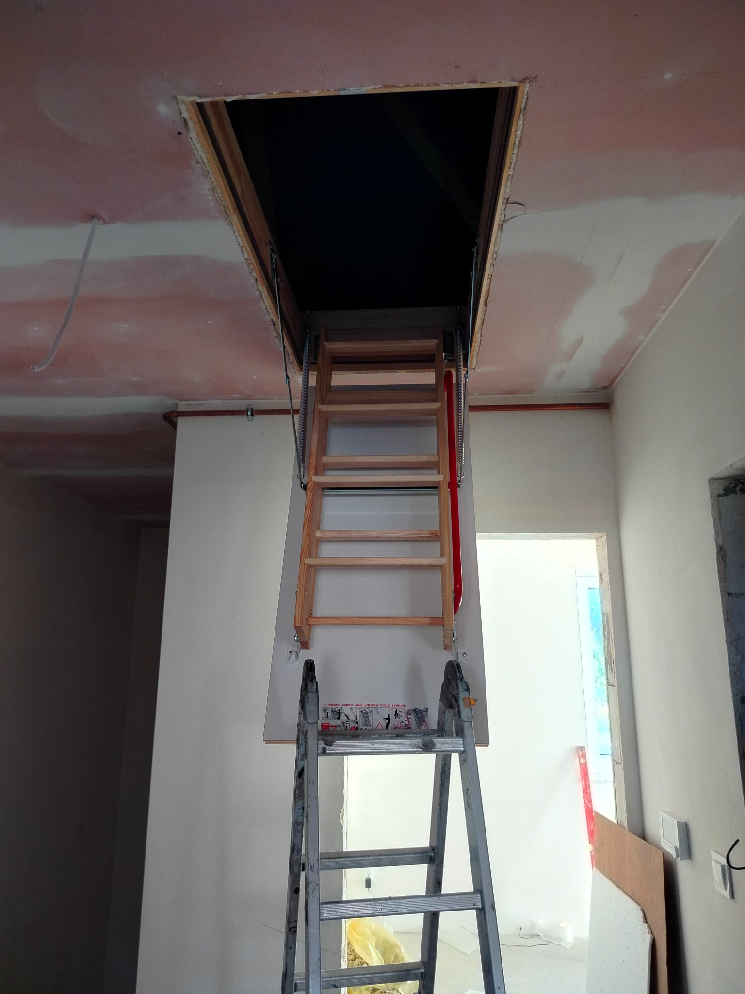 Montaż schodów strychowych: drewniane schody rozkładane z czerwoną poręczą, widoczne na tle sufitu po szpachlowaniu, z metalową drabiną pomocniczą.