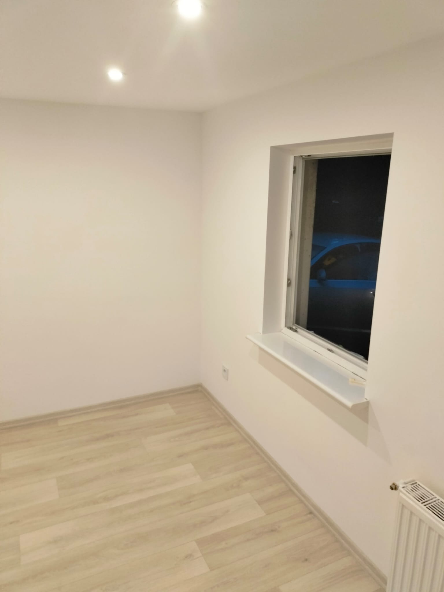 Pomalowane na biało wnętrze pokoju z jasną podłogą, oknem z widokiem na noc i białym grzejnikiem. Minimalistyczny design, świeżo po remoncie.