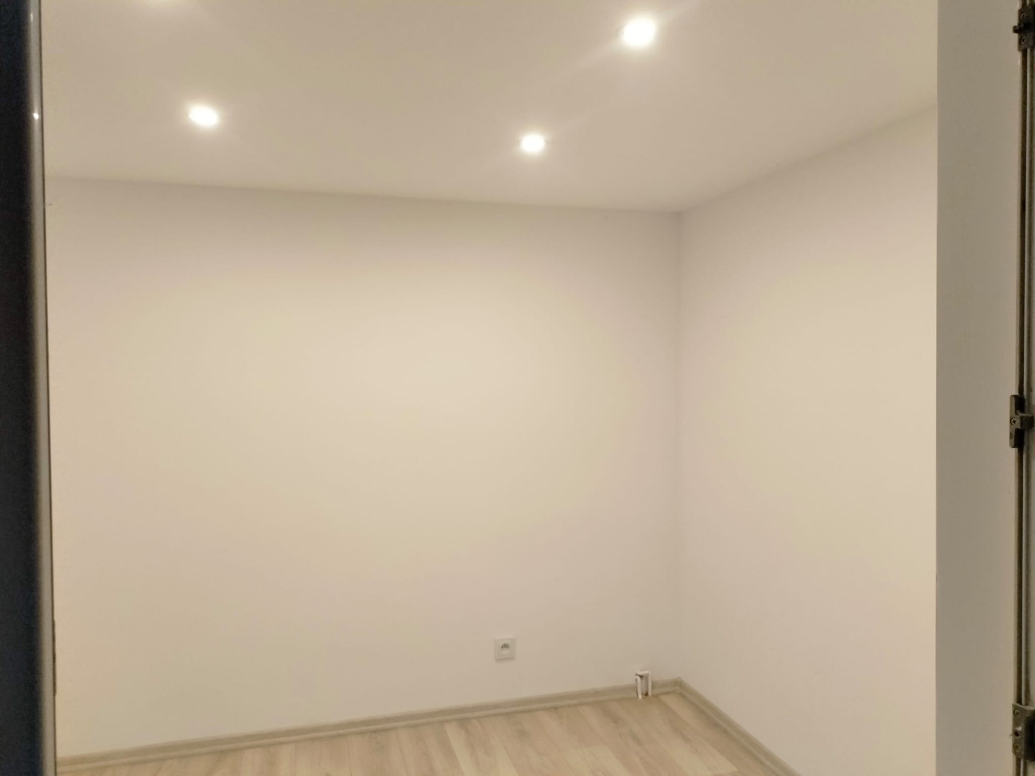 Pomalowane na biało wnętrze pokoju z trzema punktowymi lampami sufitowymi i jasną podłogą. Widoczny fragment framugi drzwi po prawej stronie kadru.
