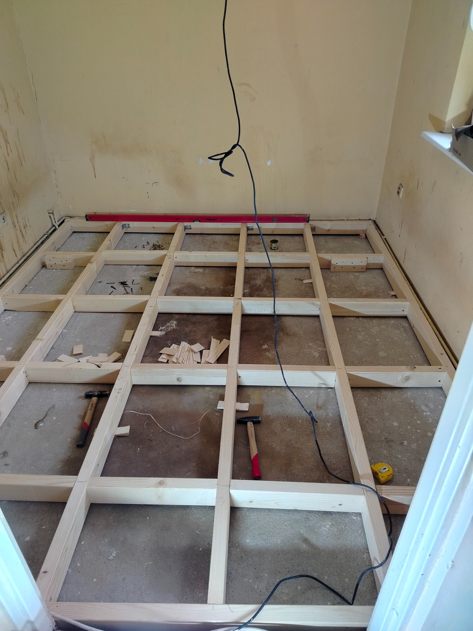 Drewniana konstrukcja pod podłogę w trakcie montażu. Widoczne poziomica, młotki i miarka. Surowe ściany w tle. Etap przygotowania podłoża pod nową podłogę.