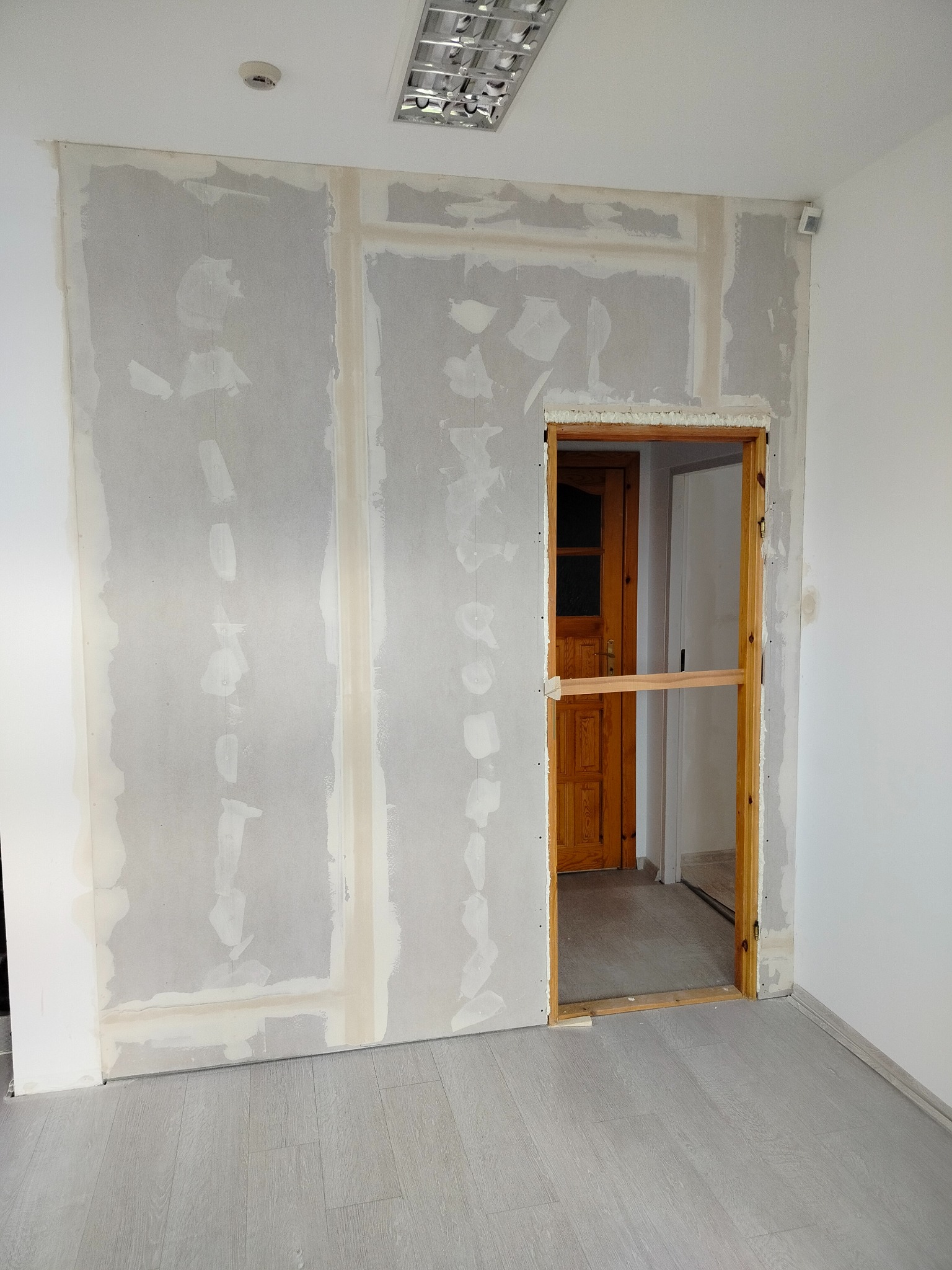 Ściana z płyt karton-gips, częściowo wyszpachlowana, z ościeżnicą drewnianą. Widoczne łączenia płyt i szpachlowane miejsca. Podłoga szara, imitująca drewno.