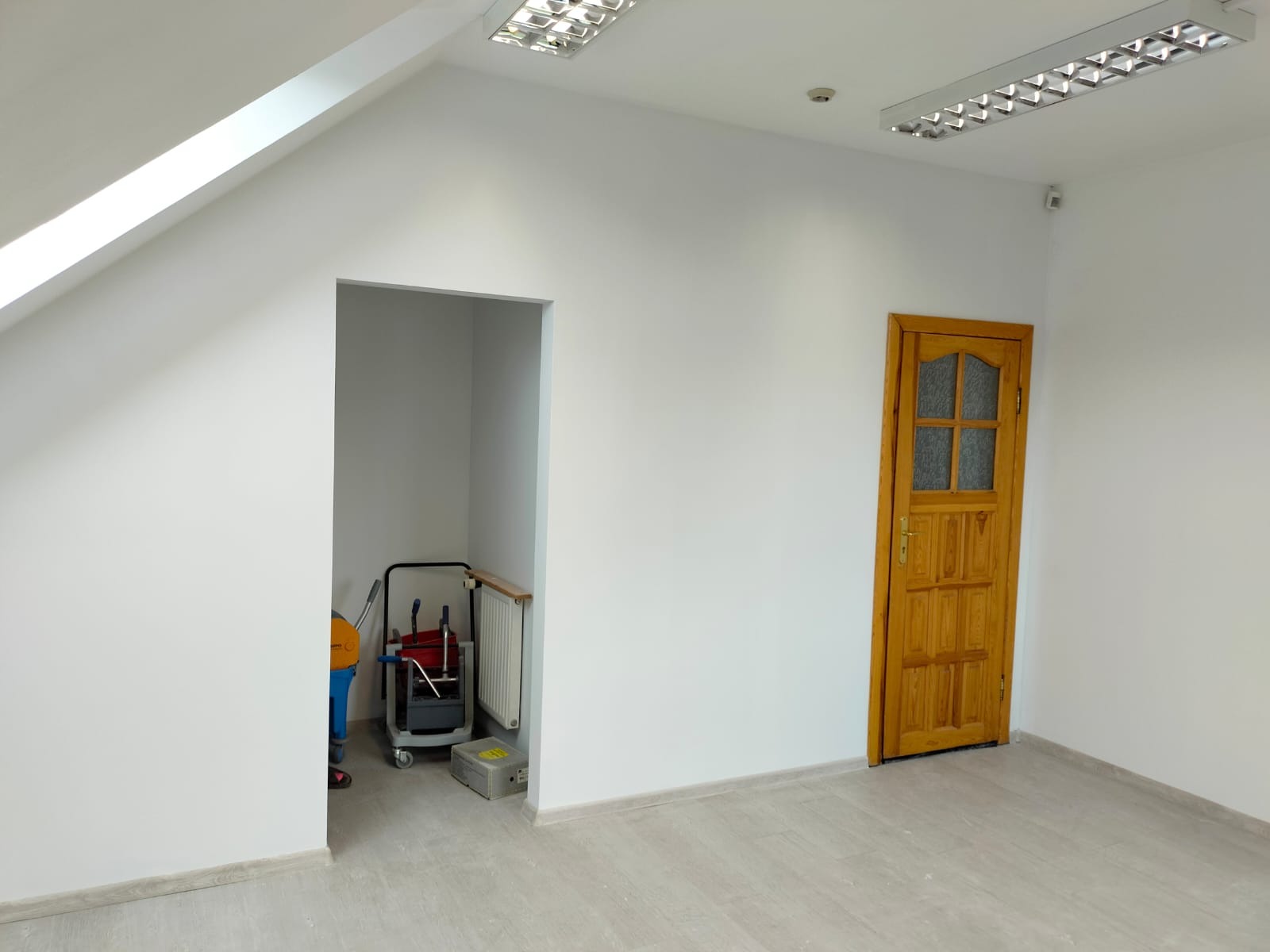 Białe, świeżo pomalowane wnętrze biura z drewnianymi drzwiami i wnęką, w której stoi wózek z narzędziami malarskimi. Widoczne oświetlenie sufitowe.