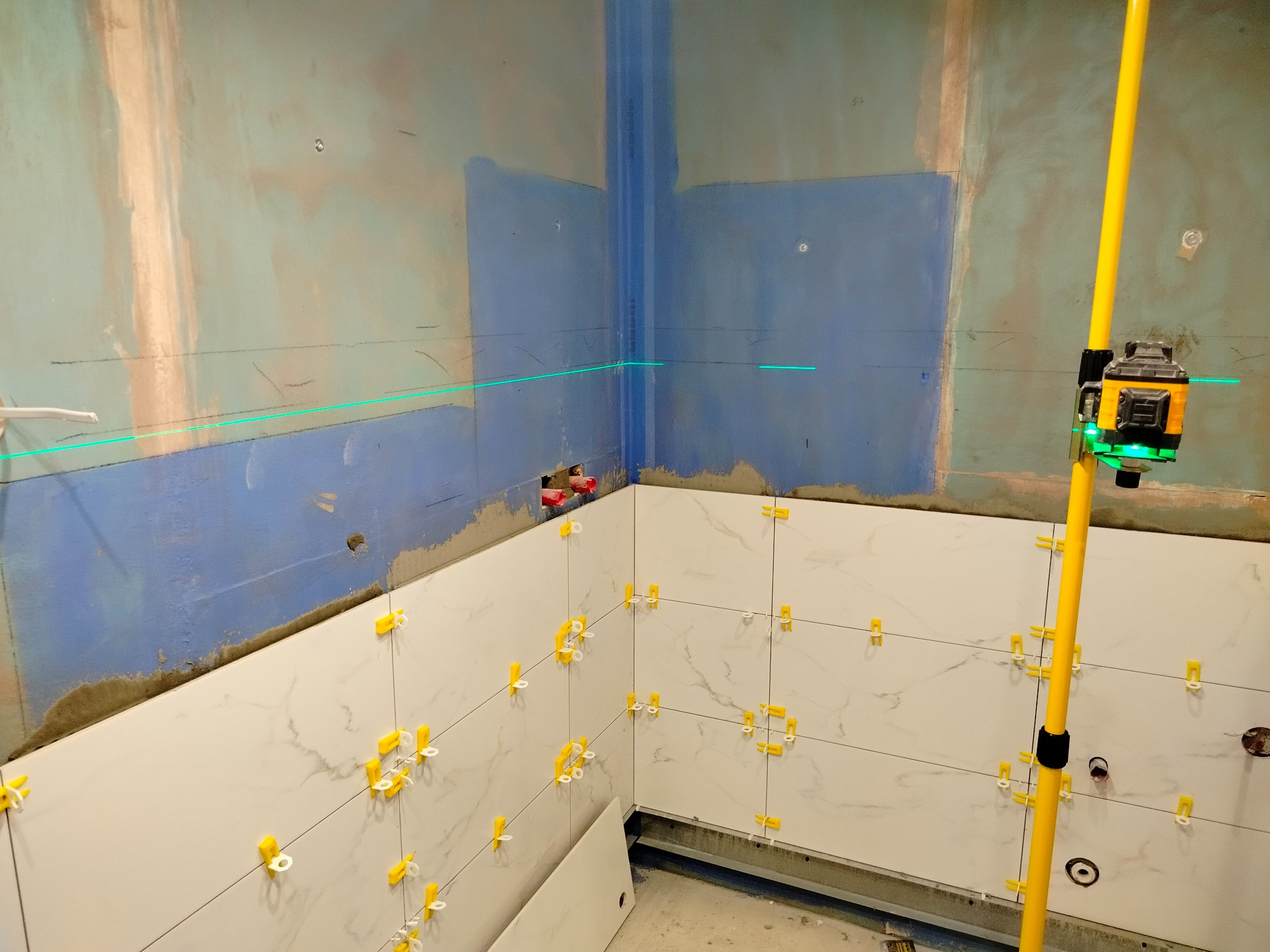Układanie białych płytek imitujących marmur w łazience, z użyciem poziomicy laserowej i żółtych krzyżyków dystansowych. Widoczne ślady po kleju na ścianach.