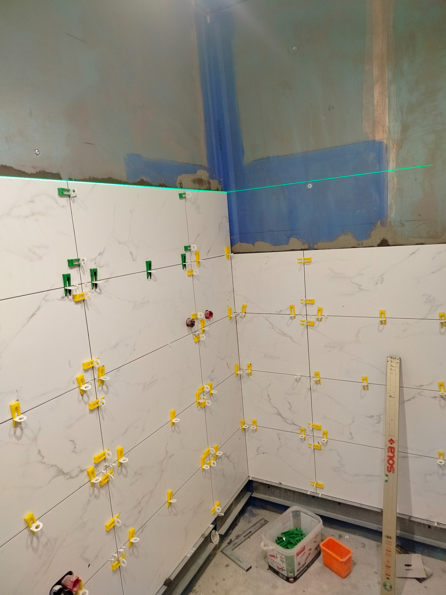 Ściana w trakcie wykańczania z białymi płytkami imitującymi marmur, poziomowana laserowo, z użyciem klipsów i klinów do glazury. Widoczne narzędzia glazurnicze.