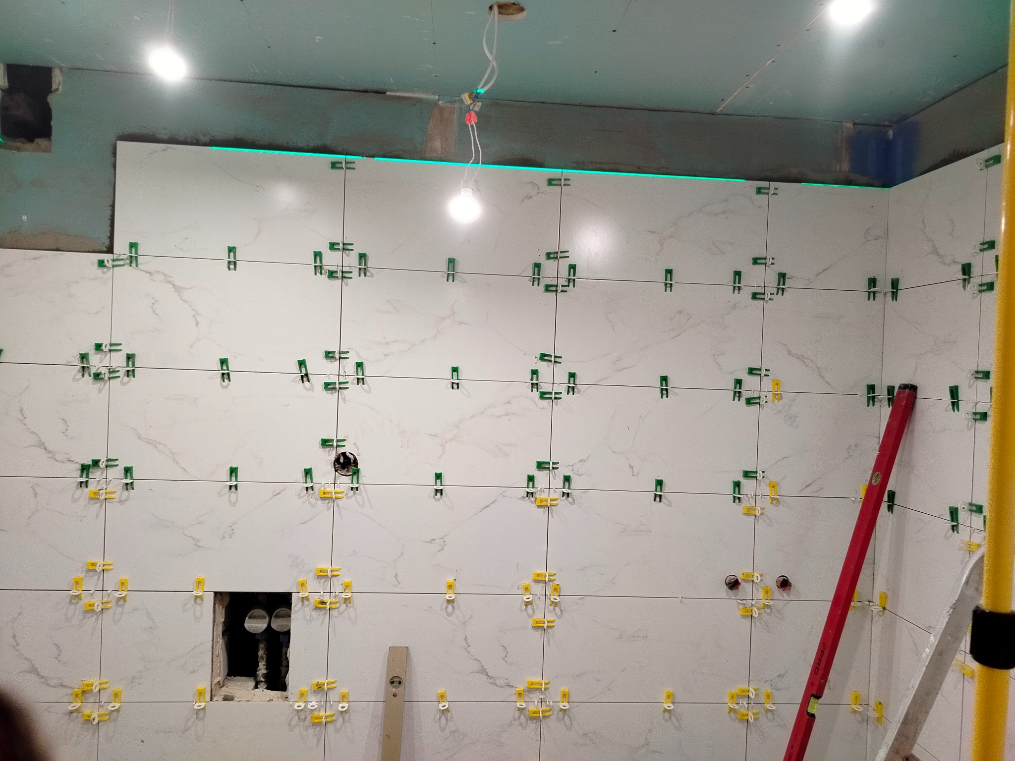 Ściana w trakcie układania białych płytek imitujących marmur, z użyciem poziomicy laserowej i klipsów poziomujących. Widoczne otwory na instalacje.