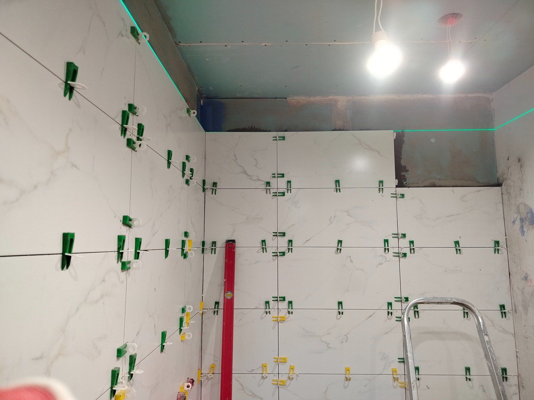 Układanie białych płytek imitujących marmur w łazience, z użyciem poziomicy laserowej i klipsów poziomujących. Widoczna drabina i fragment sufitu w trakcie prac wykończeniowych.