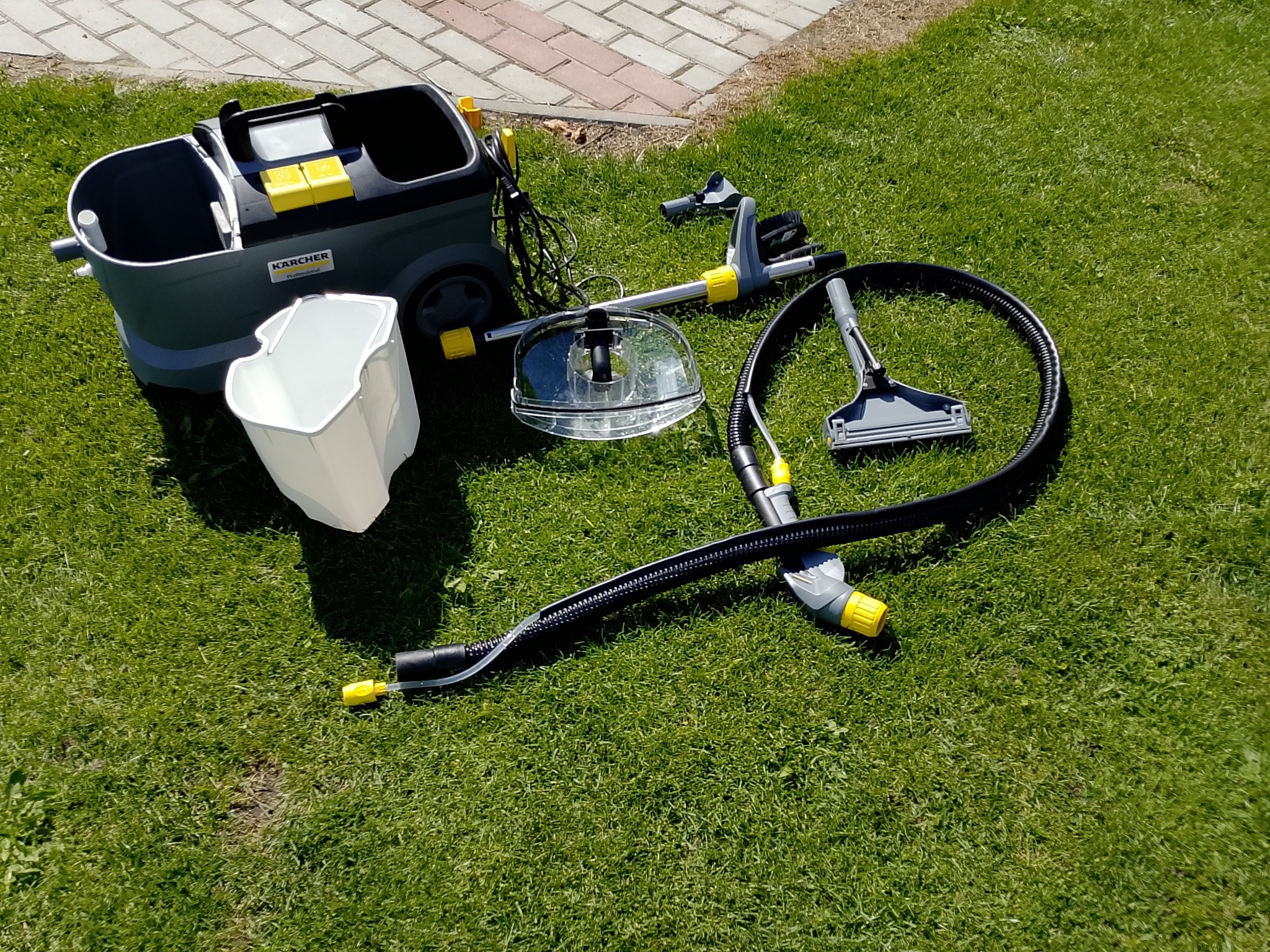 Profesjonalny odkurzacz piorący Karcher z akcesoriami na trawie w słoneczny dzień. Sprzęt do prania dywanów i tapicerki, widoczny zbiornik na wodę i różne końcówki.