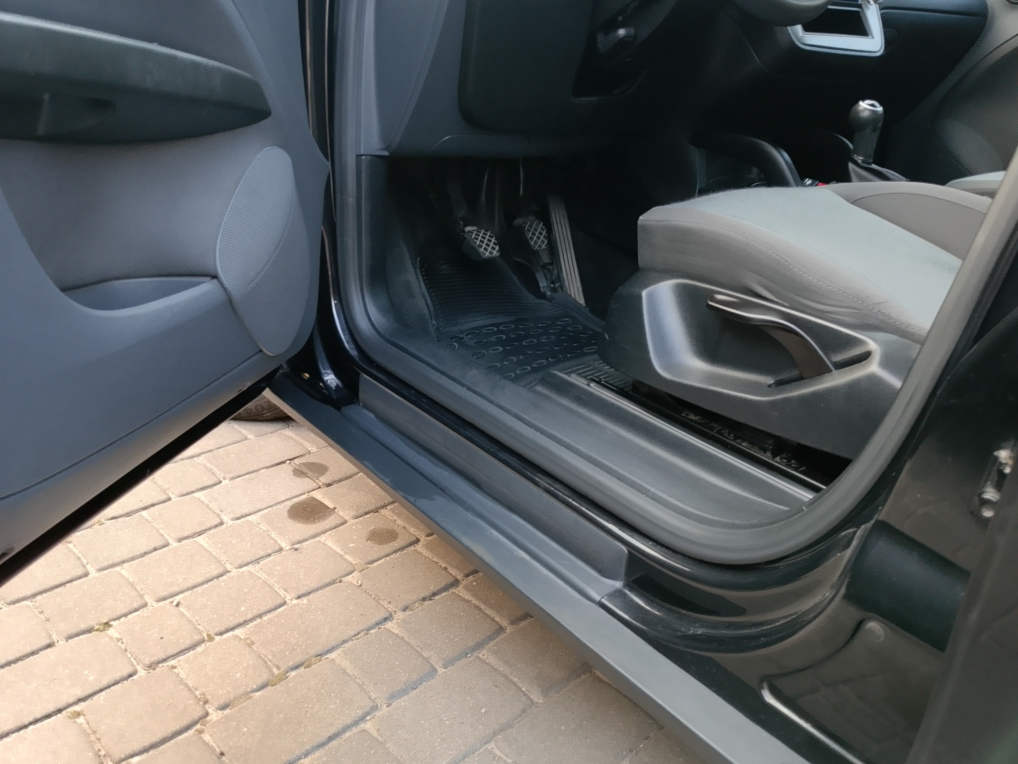 Wnętrze czarnego samochodu z otwartymi drzwiami, widoczne pedały, dywanik i część fotela na tle kostki brukowej. Ujęcie z bliska, skupione na detalu.