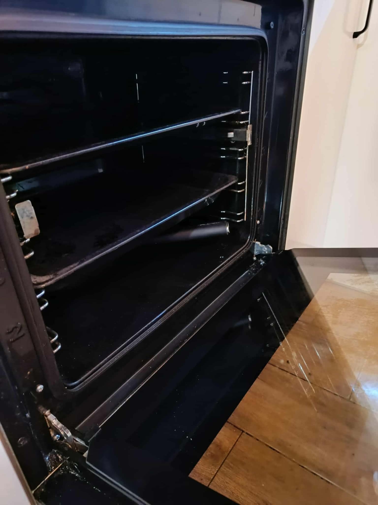 Otwarty piekarnik z widocznymi półkami i wnętrzem. Widoczne zabrudzenia na dolnej krawędzi drzwiczek i na podłodze. Ujęcie z bliska, wnętrze ciemne.