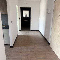 MNBwnetrza - Wykończony korytarz z panelami podłogowymi i białymi ścianami. W głębi widoczne drzwi do innego pomieszczenia z czarną futryną. W lustrze odbija się osoba.