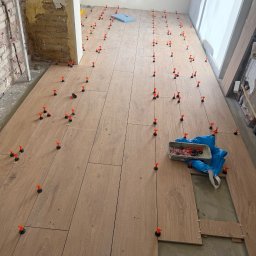 MNBwnetrza - Układanie podłogi z paneli drewnopodobnych z użyciem klipsów poziomujących. Częściowo wykończone pomieszczenie w trakcie prac. Widoczne narzędzia i materiały.