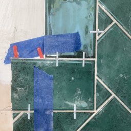 MNBwnetrza - Układanie zielonej glazury w jodełkę, zabezpieczonej niebieską taśmą i krzyżykami dystansowymi. Widoczny fragment ściany przed położeniem płytek.