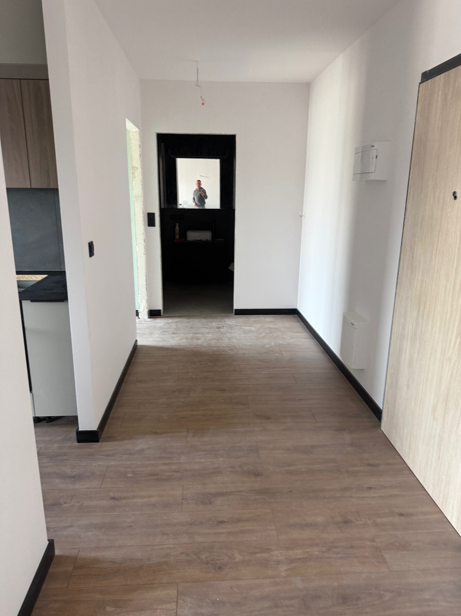 Wykończony korytarz z panelami podłogowymi i białymi ścianami. W głębi widoczne drzwi do innego pomieszczenia z czarną futryną. W lustrze odbija się osoba.