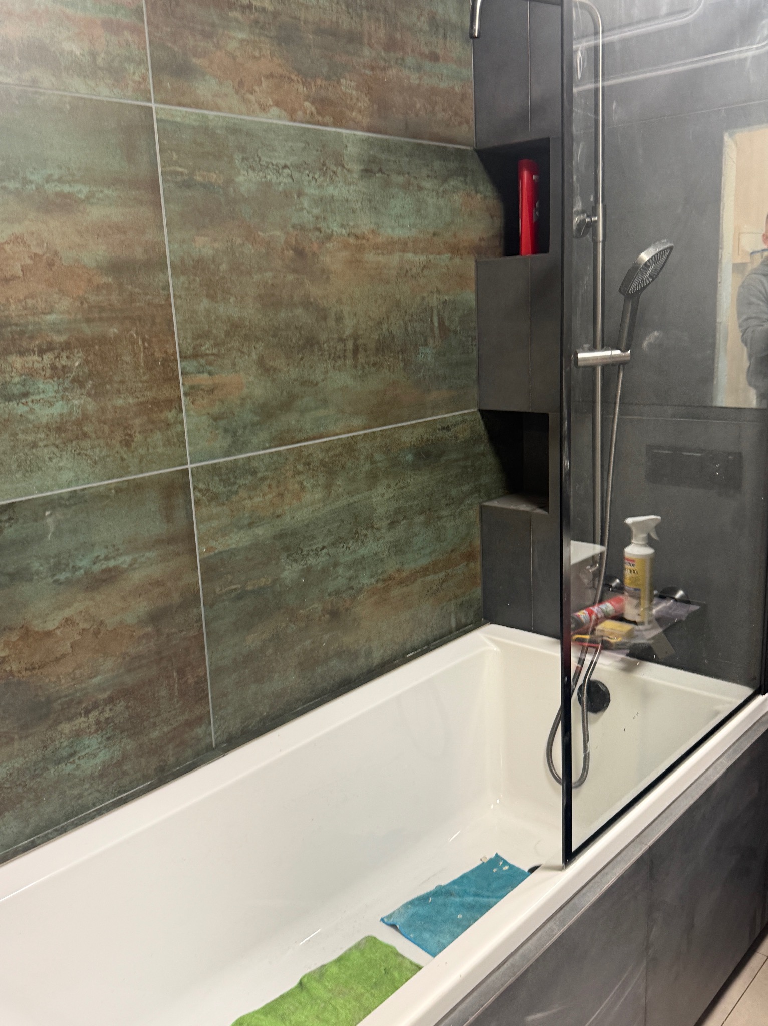 Wykończona łazienka z wanną, szklanym parawanem i oryginalnymi płytkami w odcieniach brązu i zieleni. Wnęka w ścianie z kosmetykami. Nowoczesny design.