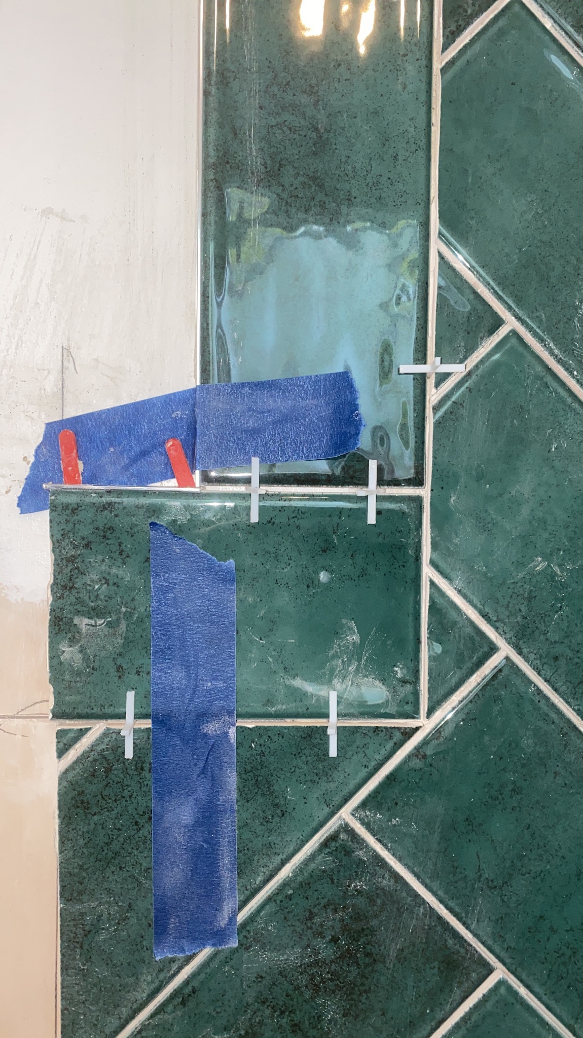 Układanie zielonej glazury w jodełkę, zabezpieczonej niebieską taśmą i krzyżykami dystansowymi. Widoczny fragment ściany przed położeniem płytek.