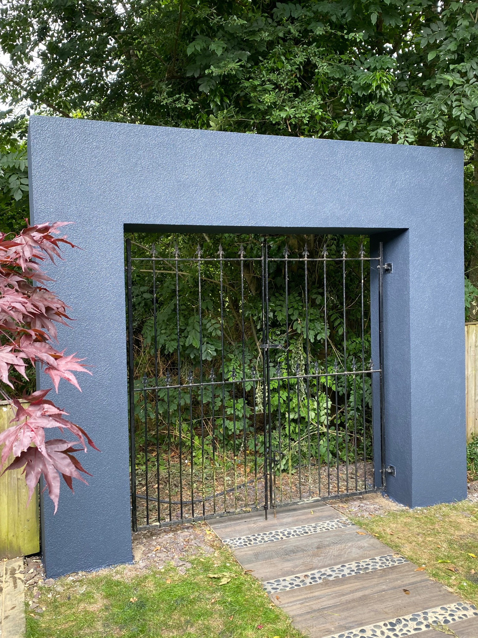 Szara, otynkowana rama z metalową bramą w ogrodzie. Widoczne drewniane ścieżki i zieleń. Świeżo pomalowana konstrukcja z bramą.