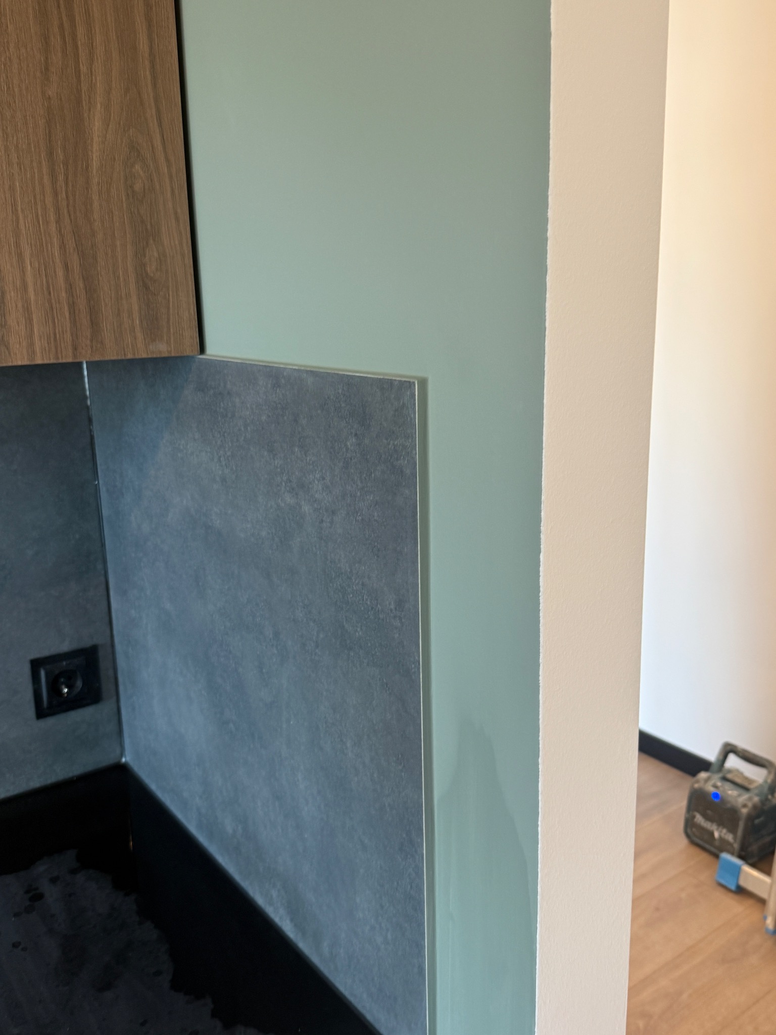 Fragment wykończonej kuchni: szara ściana z płytkami, drewniana szafka, świeżo pomalowana ściana na pistacjowo, narzędzia malarskie w tle.