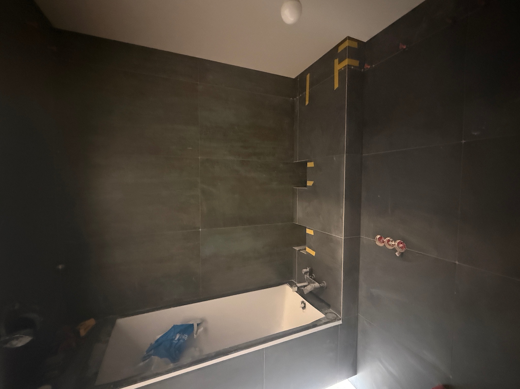 Ciemne płytki w łazience w trakcie montażu, widoczna wanna zabezpieczona folią, taśma malarska na fugach i wystające przyłącza wodne.