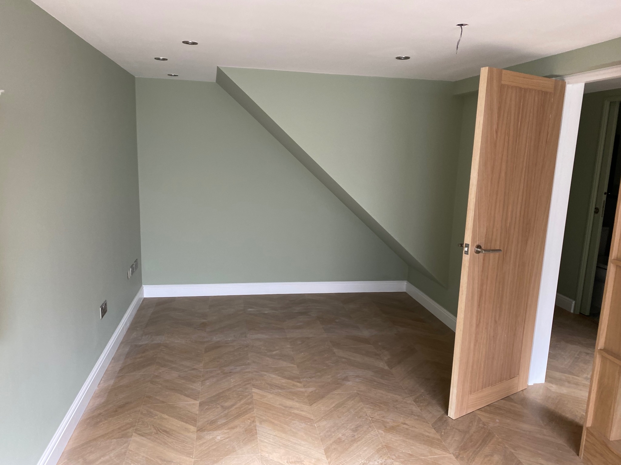 Jasne wnętrze po remoncie: zielone ściany, parkiet w jodełkę, białe listwy przypodłogowe i drewniane drzwi z klamką. Widoczne gniazdka elektryczne i oświetlenie sufitowe.