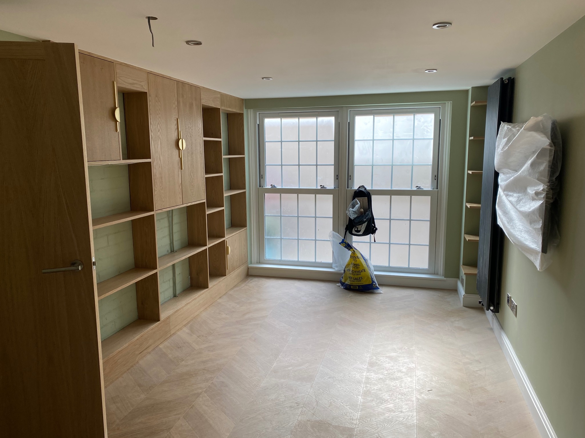 Jasne, drewniane regały z półkami i szafkami z złotymi uchwytami, ułożona podłoga w jodełkę, okno z widokiem na zewnątrz, ściany w odcieniach zieleni.