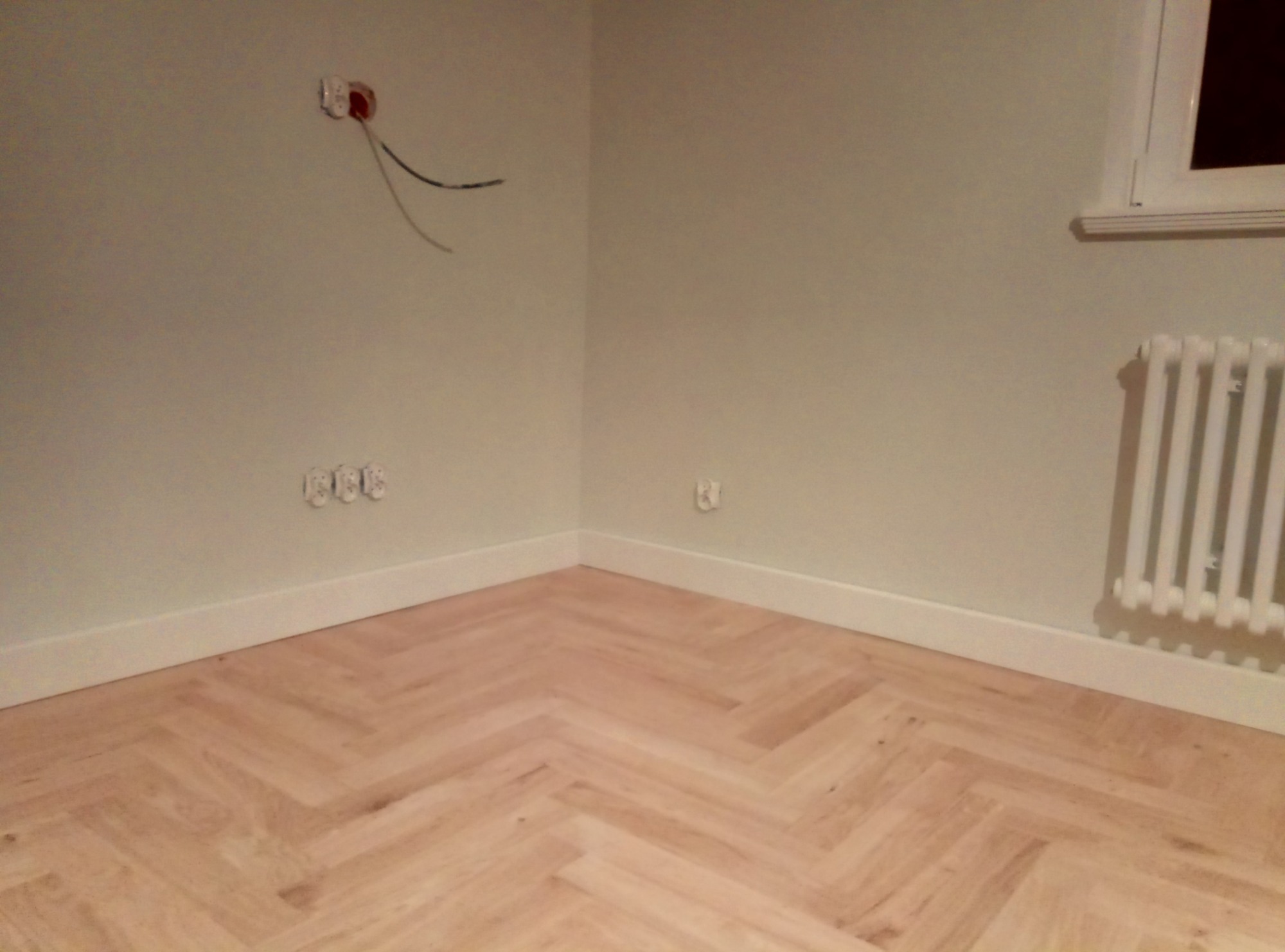 Jasny parkiet ułożony w jodełkę w rogu pokoju z grzejnikiem i gniazdkami. Ściany w odcieniach bieli, widoczne przewody elektryczne.