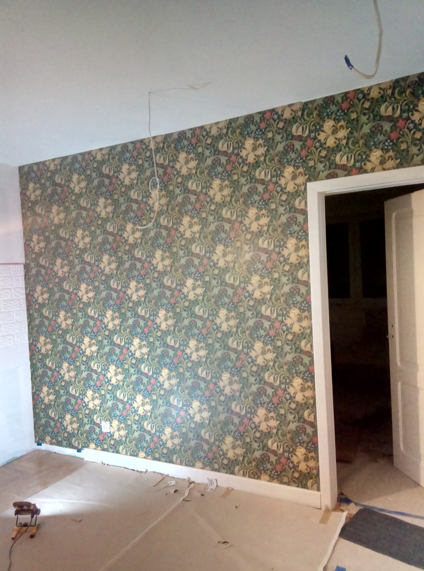 Pokój po tapetowaniu ścian wzorzystą tapetą w stylu vintage. Widoczne niedociągnięcia przy podłodze i wystające kable. Otwór drzwiowy z białą ościeżnicą.
